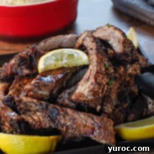 Greek ribs piled on a black serving plate with lemon wedges and a red dish of rice in and grey oven mitts in the background