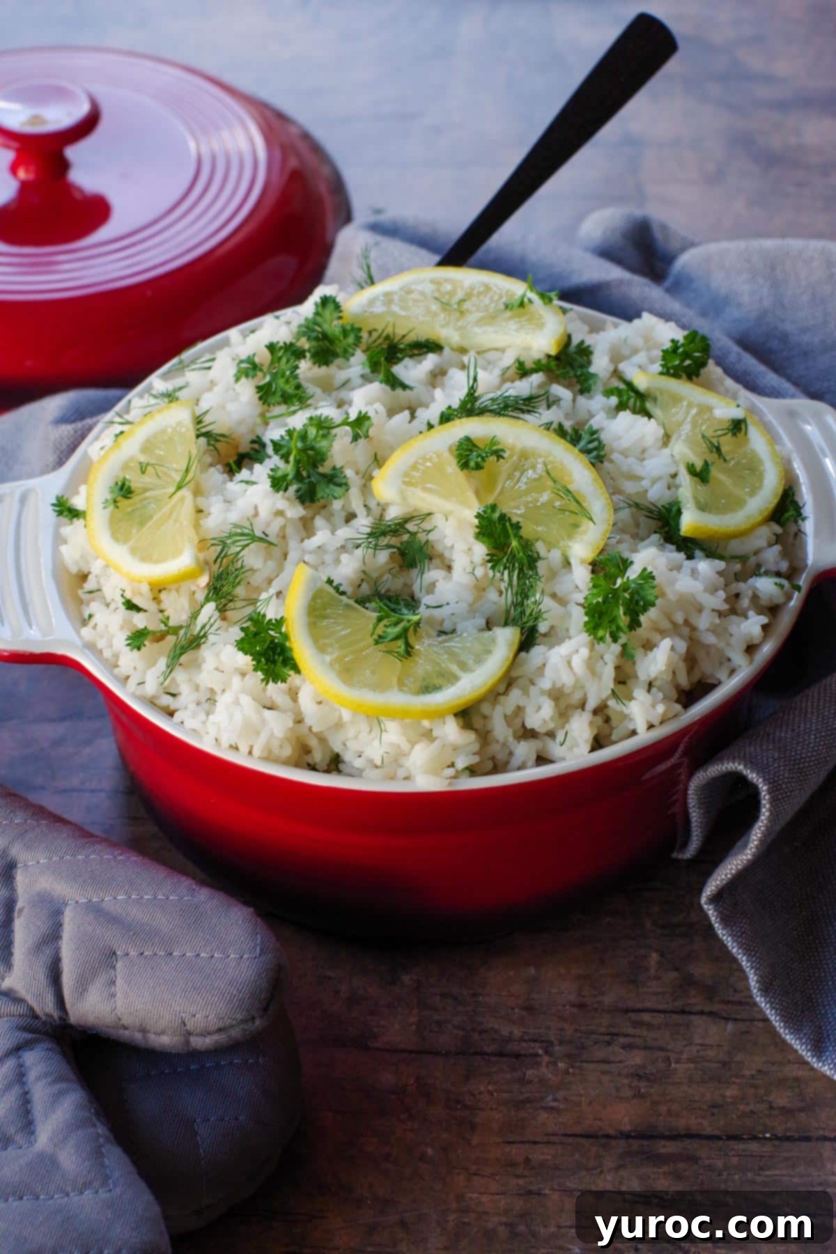 Greek rice with lemon and herb garnish in orange dish with orange lid in the background and grey oven mitt on the left side