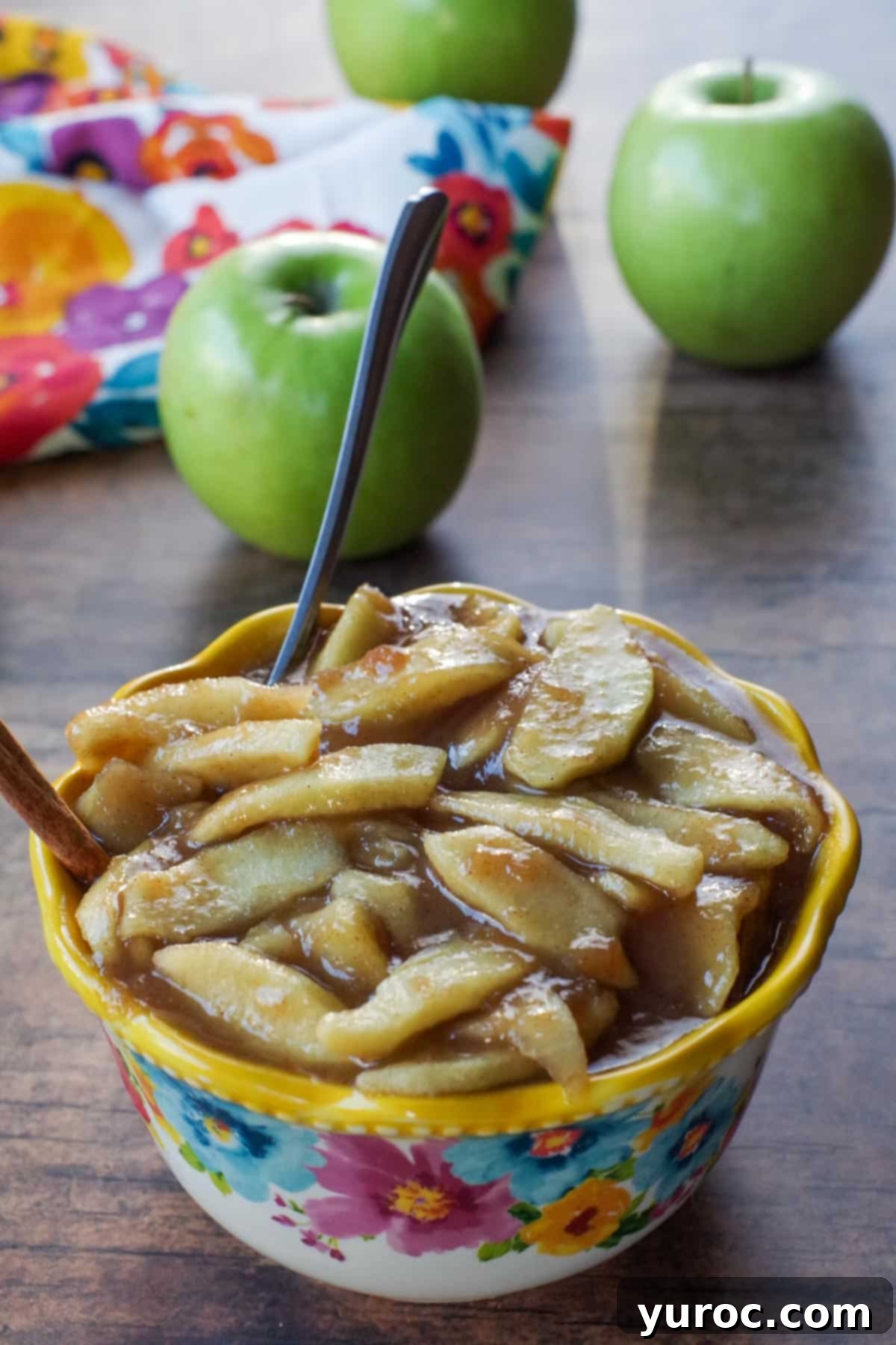 stovetop apple pie filling in a flowered blue bowl with yellow rim, with spoon and cinnamon stick in it and green apples in background