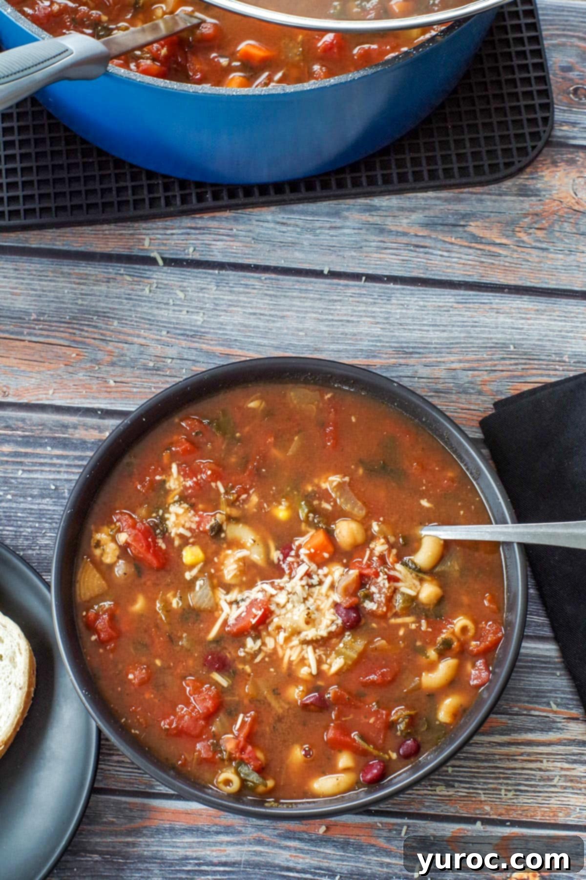 Wholesome Minestrone Made Easy 9 A black bowl brimming with minestrone soup, featuring a spoon resting in it. In the soft-focused background, a blue pot holds more soup, hinting at a large, comforting batch.