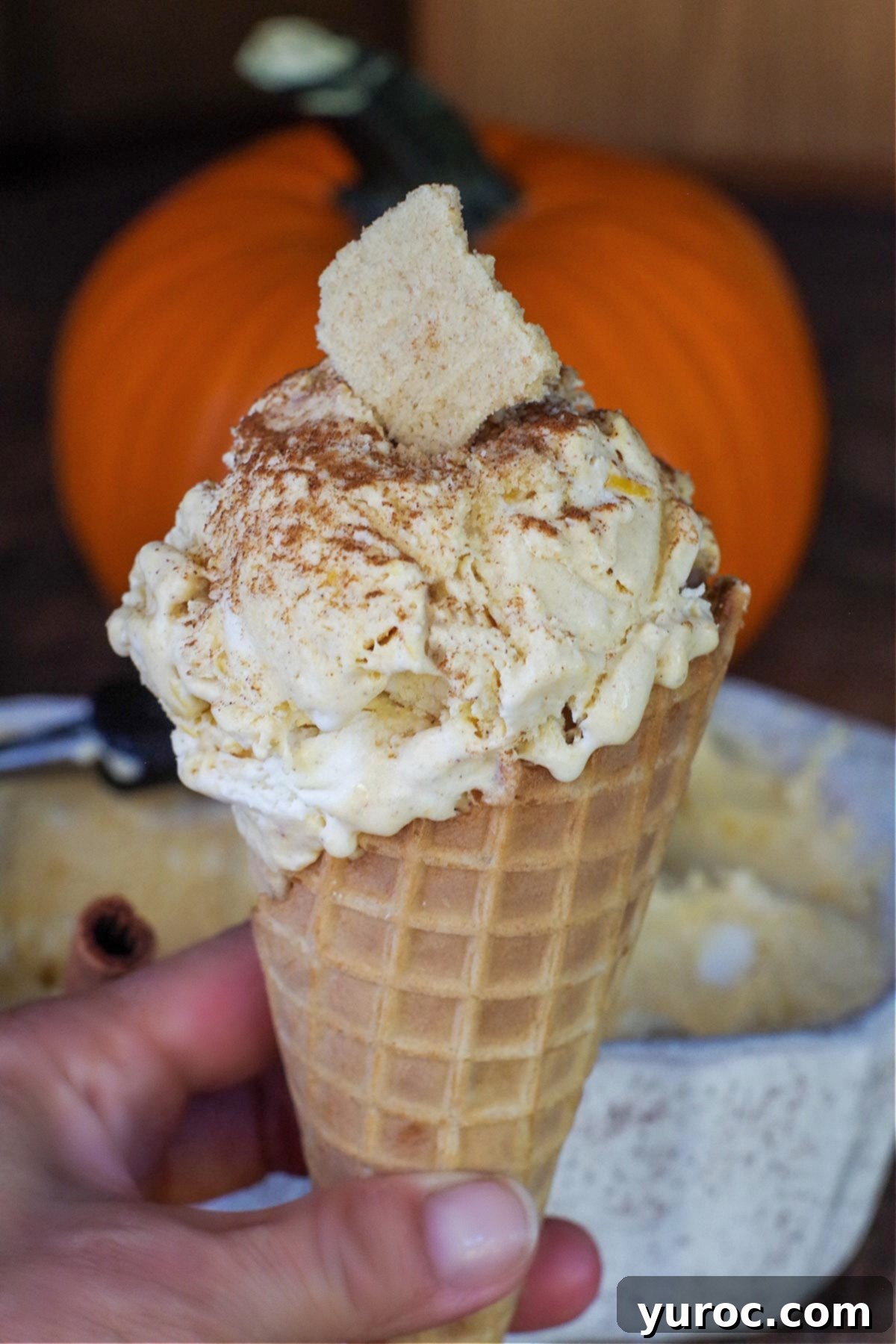 A scoop of homemade no-churn pumpkin pie ice cream in a sugar cone, held by a hand. A container of ice cream and a small decorative pumpkin are visible in the background.
