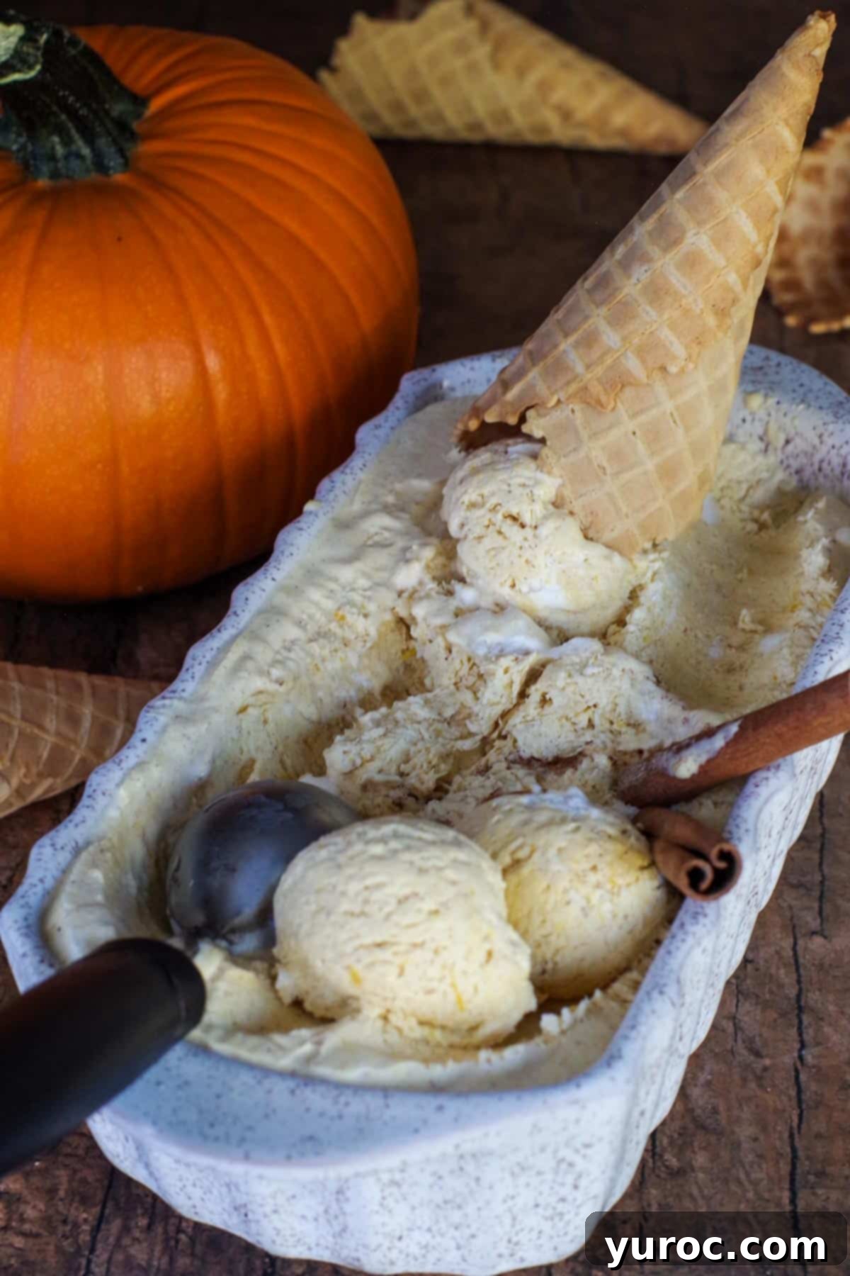 A beige ceramic loaf container filled with scooped pumpkin ice cream. An ice cream scoop, cinnamon sticks, and an empty sugar cone are arranged alongside the container.