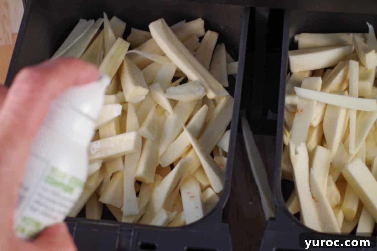 Uncooked parsnip fries in an air fryer drawer being sprayed with cooking oil, ready for seasoning.