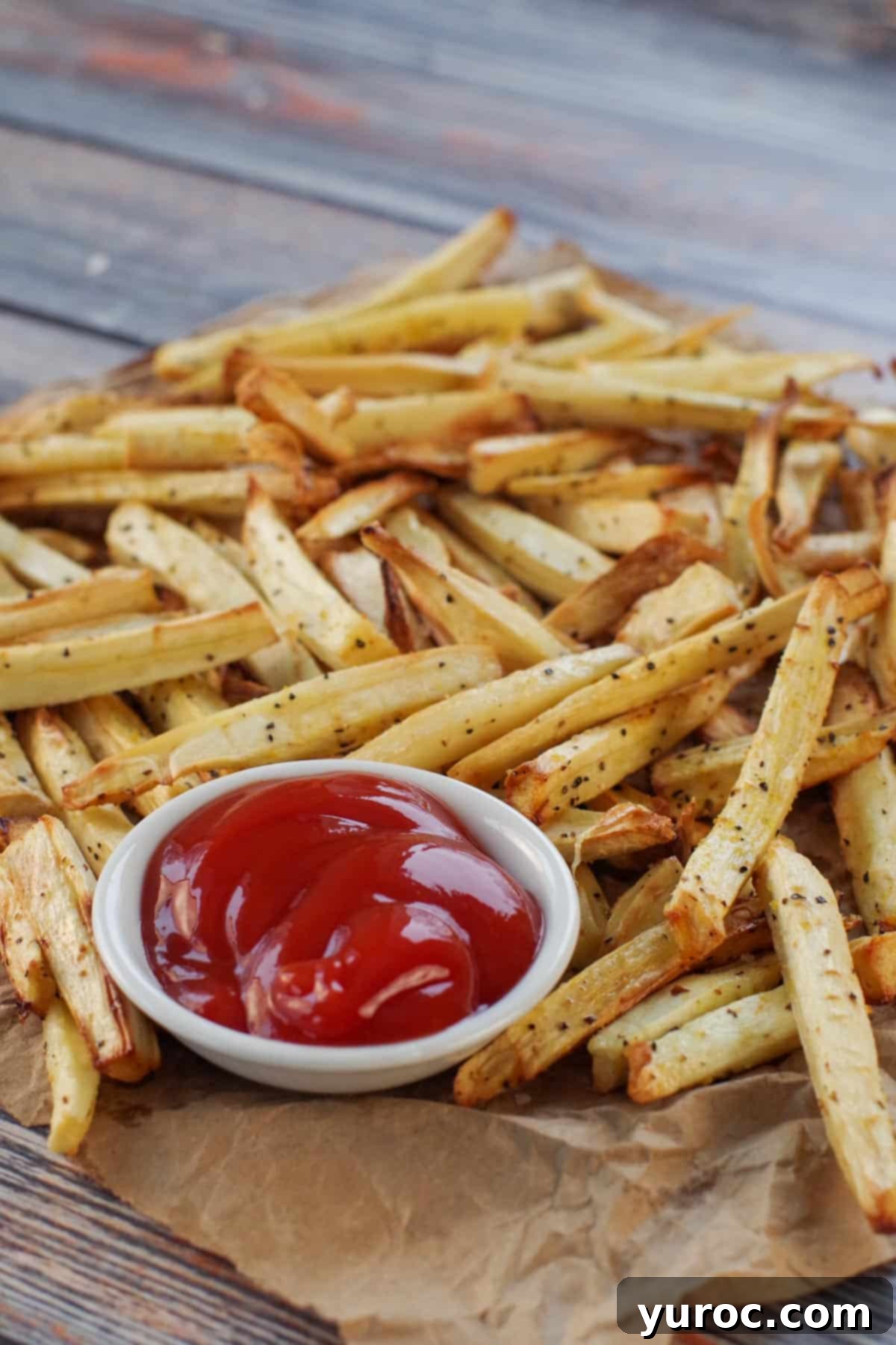 A generous serving of crispy air fryer parsnip fries resting on brown paper, with a small white bowl of ketchup in the foreground.