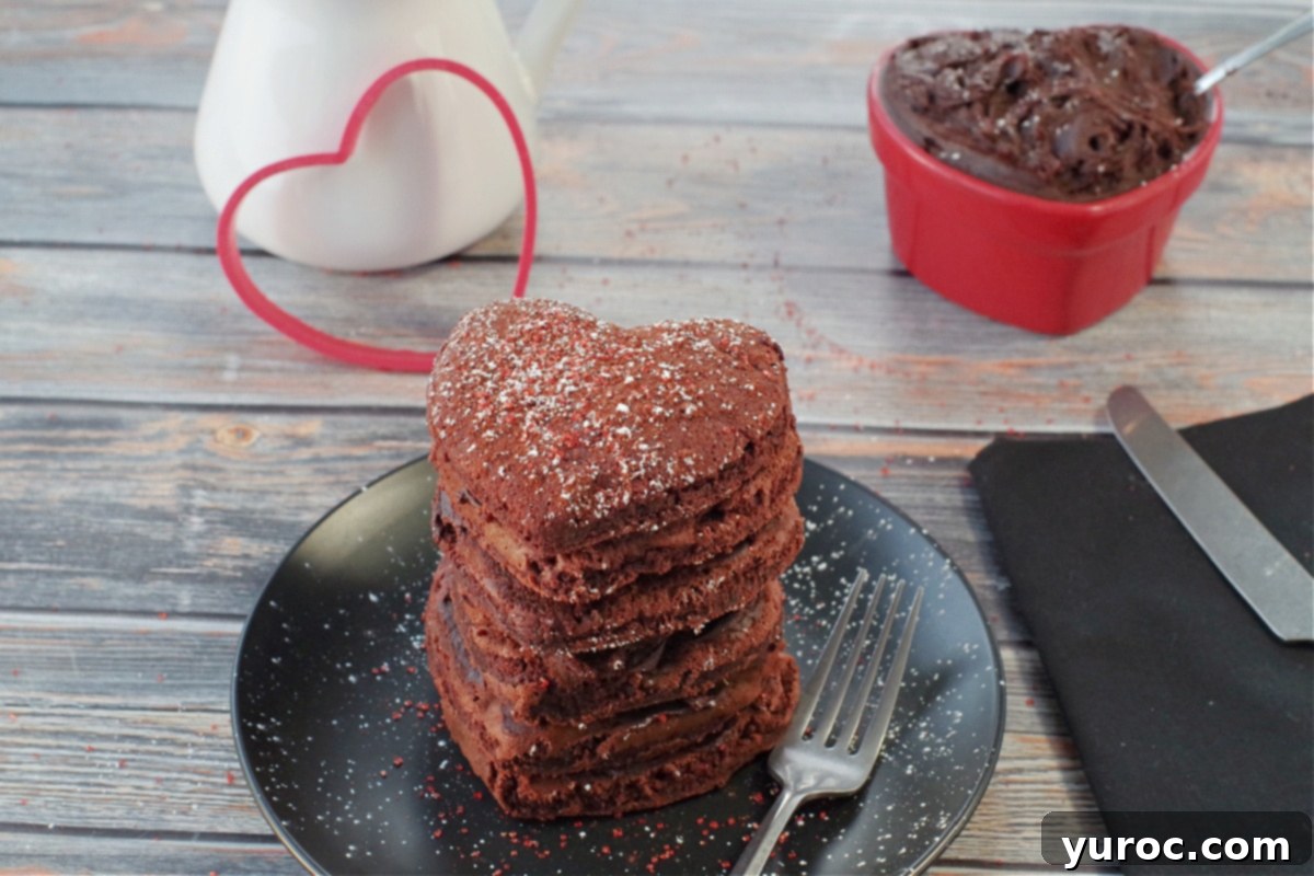 Healthy Red Velvet Pancakes with Luscious Chocolate Cream Cheese Butter 2 Stack of six healthy red velvet pancakes on a black plate with a fork, a heart-shaped dish of chocolate cream cheese butter, and a syrup container in the background.