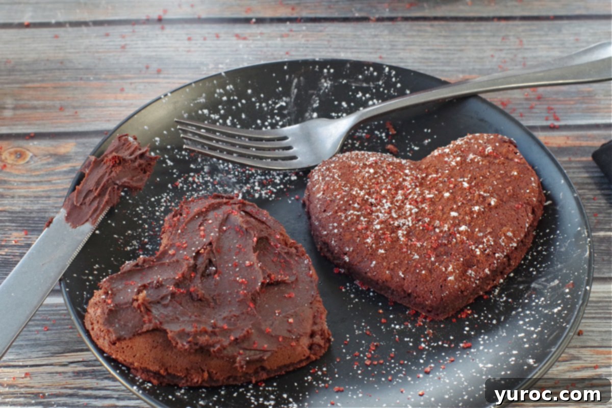 Healthy Red Velvet Pancakes with Luscious Chocolate Cream Cheese Butter 3 Two heart-shaped red velvet pancakes on a black plate, one topped with chocolate cream cheese butter and the other with red sparkling sugar and powdered sugar.