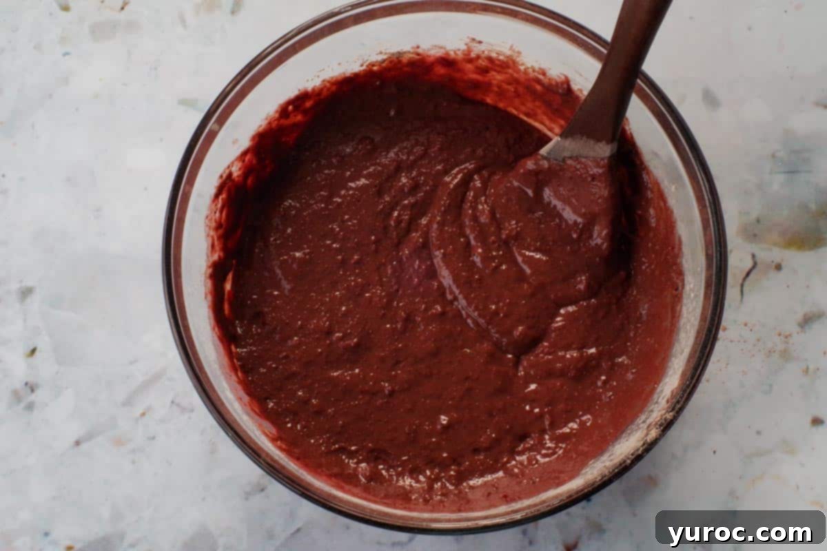 Healthy Red Velvet Pancakes with Luscious Chocolate Cream Cheese Butter 10 Wet and dry ingredients combined in a large glass bowl, mixed with a dark brown wooden spoon, showing a slightly lumpy batter.
