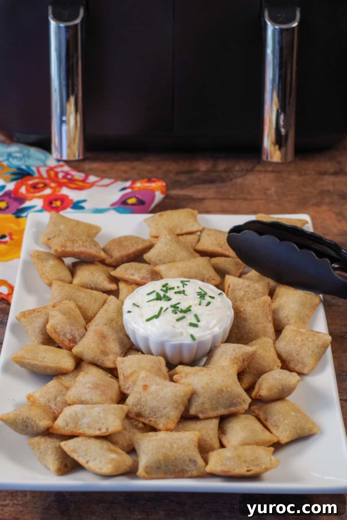 Air fryer pizza bites arranged on a white serving tray with a bowl of dipping sauce and silicone-tipped tongs. An air fryer is visible in the background.
