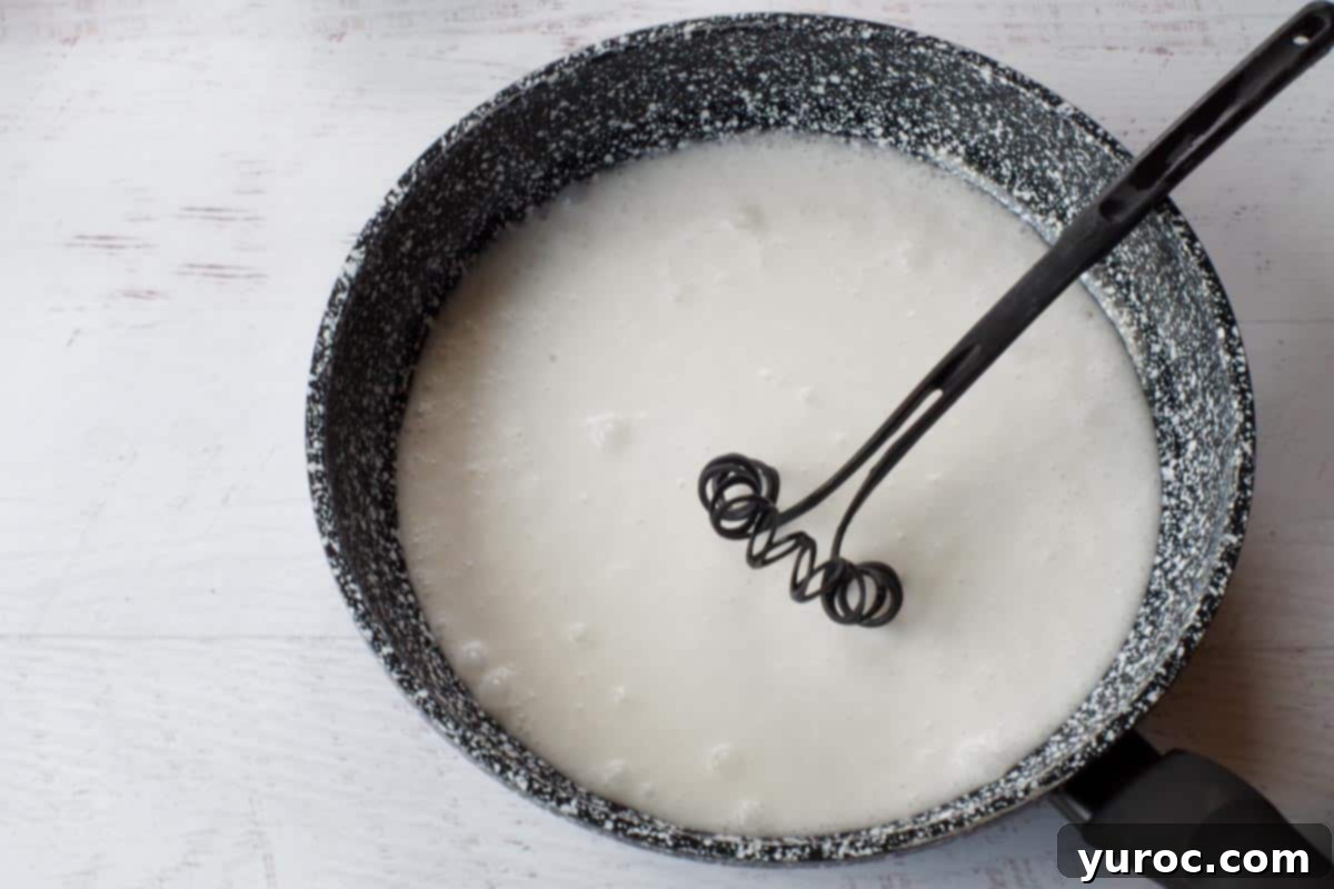 Light coconut milk being gently heated in a large, deep frying pan on the stovetop, stirred with a whisk until warmed through.