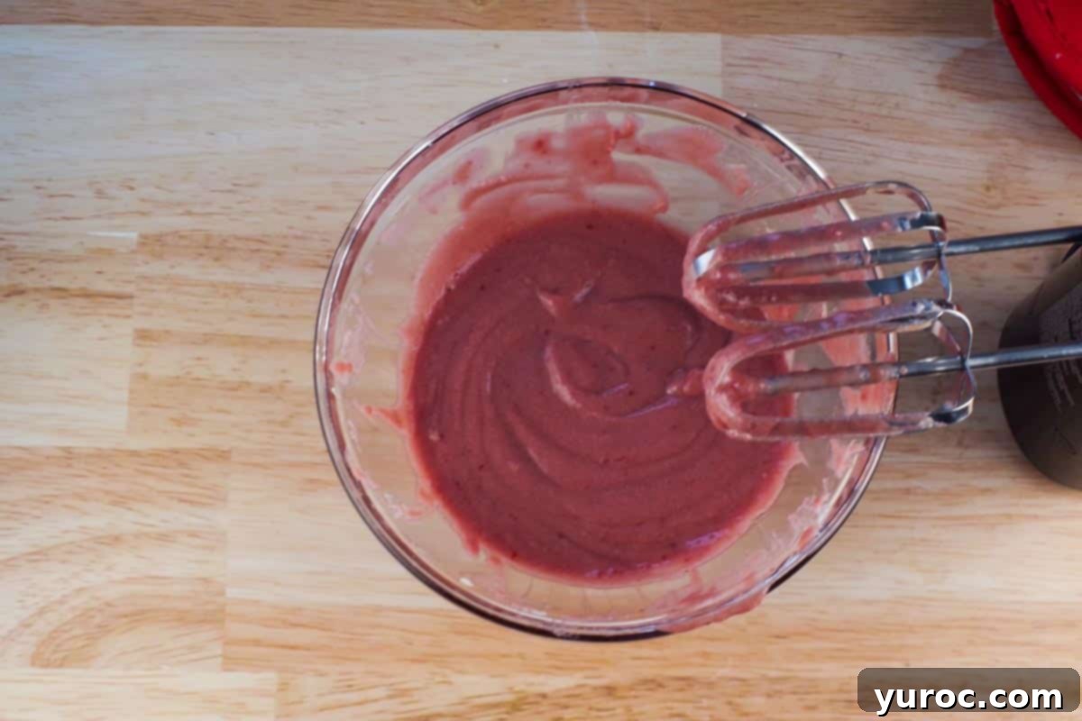 Seedless raspberry jam beaten with mascarpone cheese in a glass bowl, with beaters above.