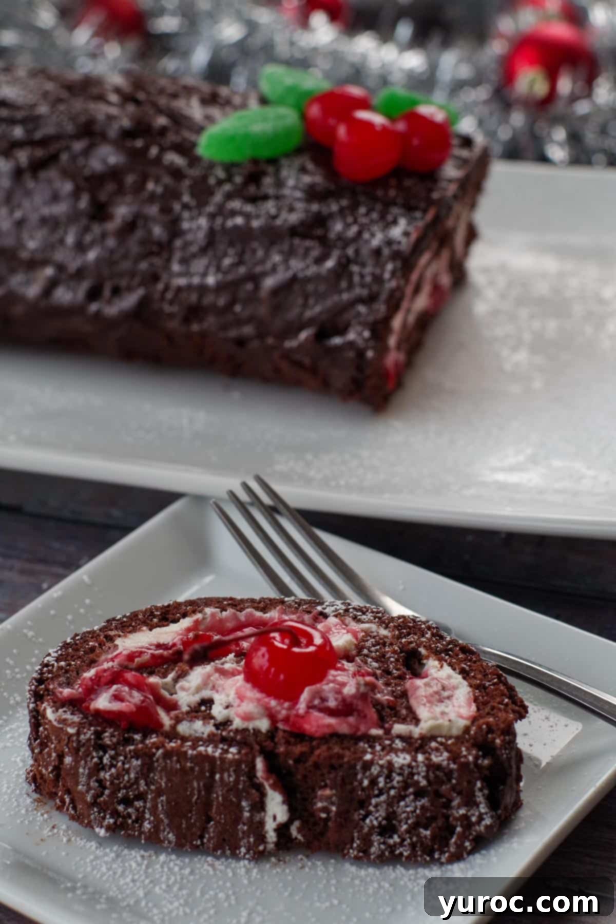 slice of black forest yule log cake on a white square plate with a fork and a whole black forest yule log cake in the background