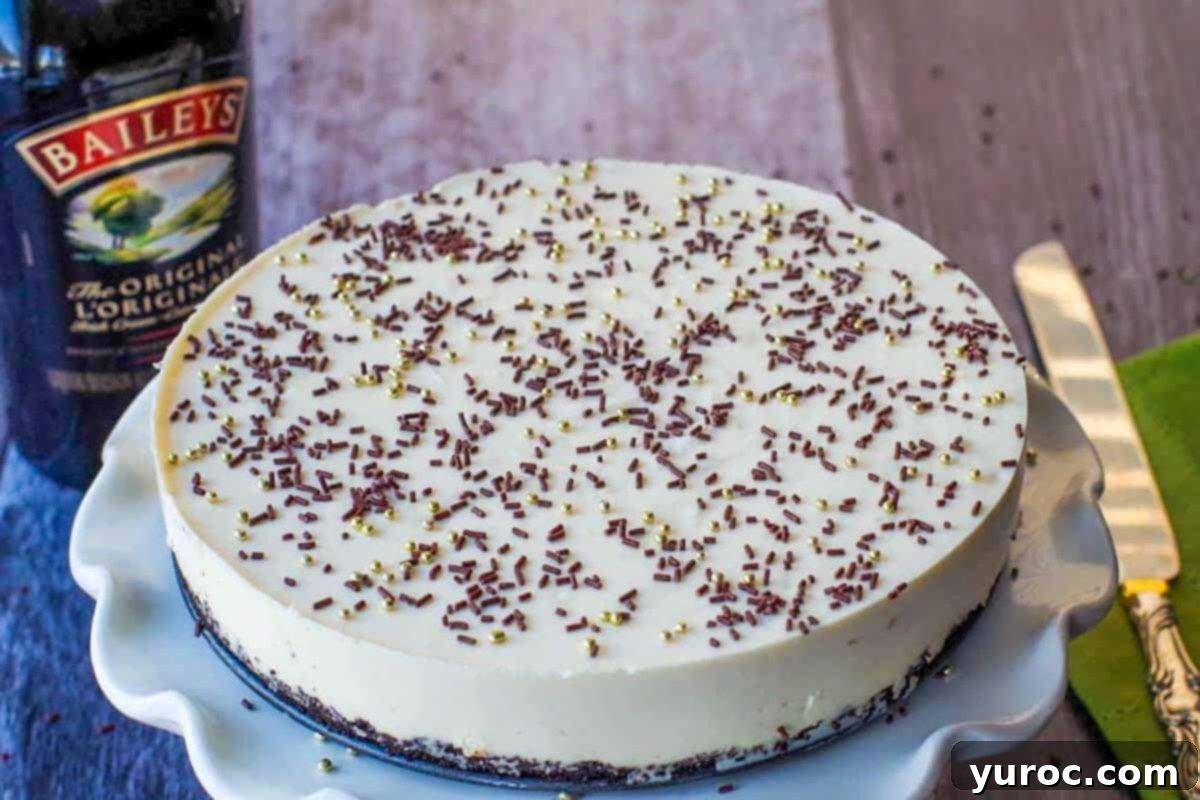 Whole No bake Baileys cheesecake on a white cake platter with a bottle of baileys in the background on the left and a knife on the right
