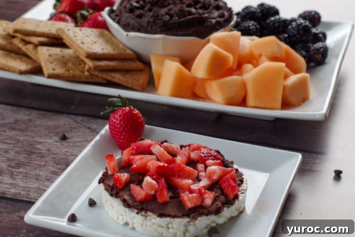 Decadent Dark Chocolate Date Hummus 11 A rice cake generously spread with dark chocolate hummus and topped with fresh strawberry slices. In the background, a platter of the hummus with fruit and graham crackers hints at more delicious options.