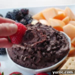 Decadent Dark Chocolate Date Hummus 13 A top-down view of creamy dark chocolate hummus in a elegant white petal bowl, nestled on a white platter with an assortment of fresh fruit and crunchy crackers. A juicy strawberry is being dipped into the hummus, emphasizing its smooth texture.