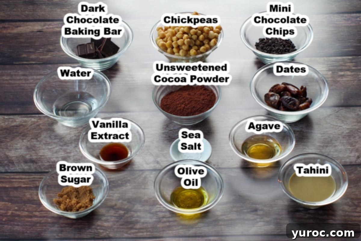 Decadent Dark Chocolate Date Hummus 3 Various ingredients for dark chocolate hummus laid out in clear glass bowls on a white surface, neatly labeled for easy identification. These include chickpeas, cocoa powder, dates, chocolate bar, and chocolate chips.