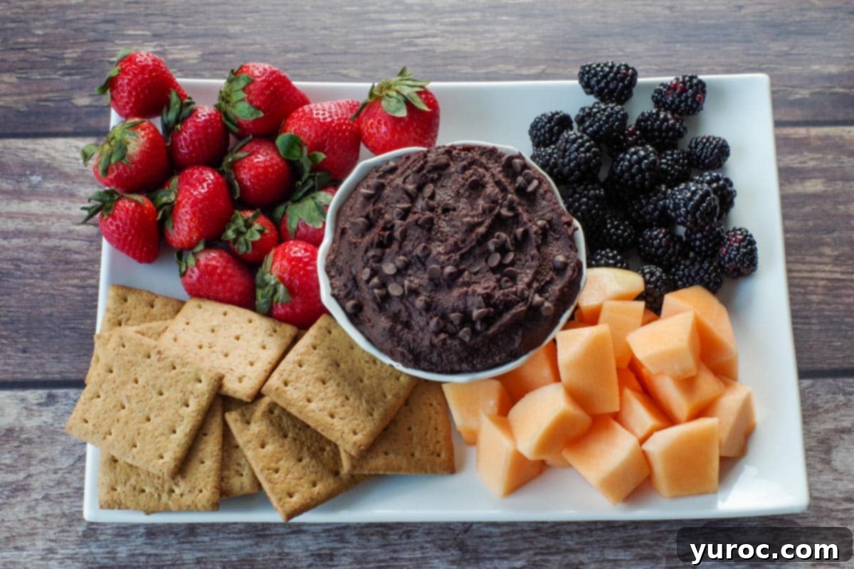 Decadent Dark Chocolate Date Hummus 9 A vibrant platter featuring a white petal bowl of creamy chocolate dessert hummus surrounded by fresh fruit like strawberries and blueberries, and crunchy graham crackers, ready for serving.