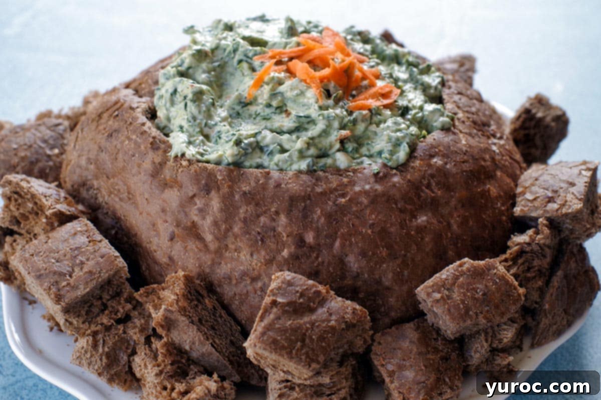 Ultimate Pumpernickel Spinach Dip 8 Pumpernickel spinach dip served in a bread bowl, surrounded by cubed bread pieces for dipping on a white plate.