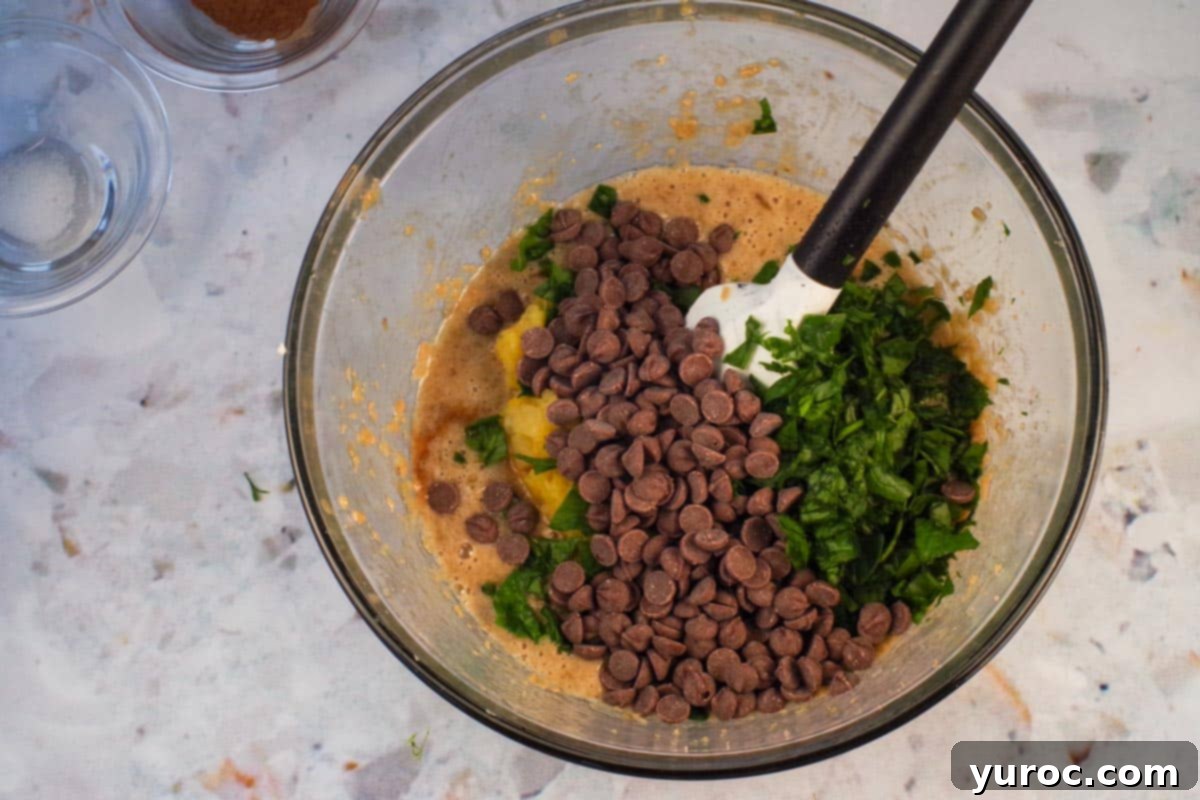 Spinach,, pineapple and chocolate chips added to mixture in large glass bowl with spatula.