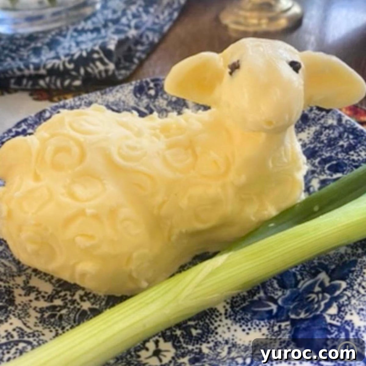 An exquisitely carved lamb figure made entirely of butter, positioned delicately on a blue floral patterned fabric, with a thin slice of green onion thoughtfully placed in front.