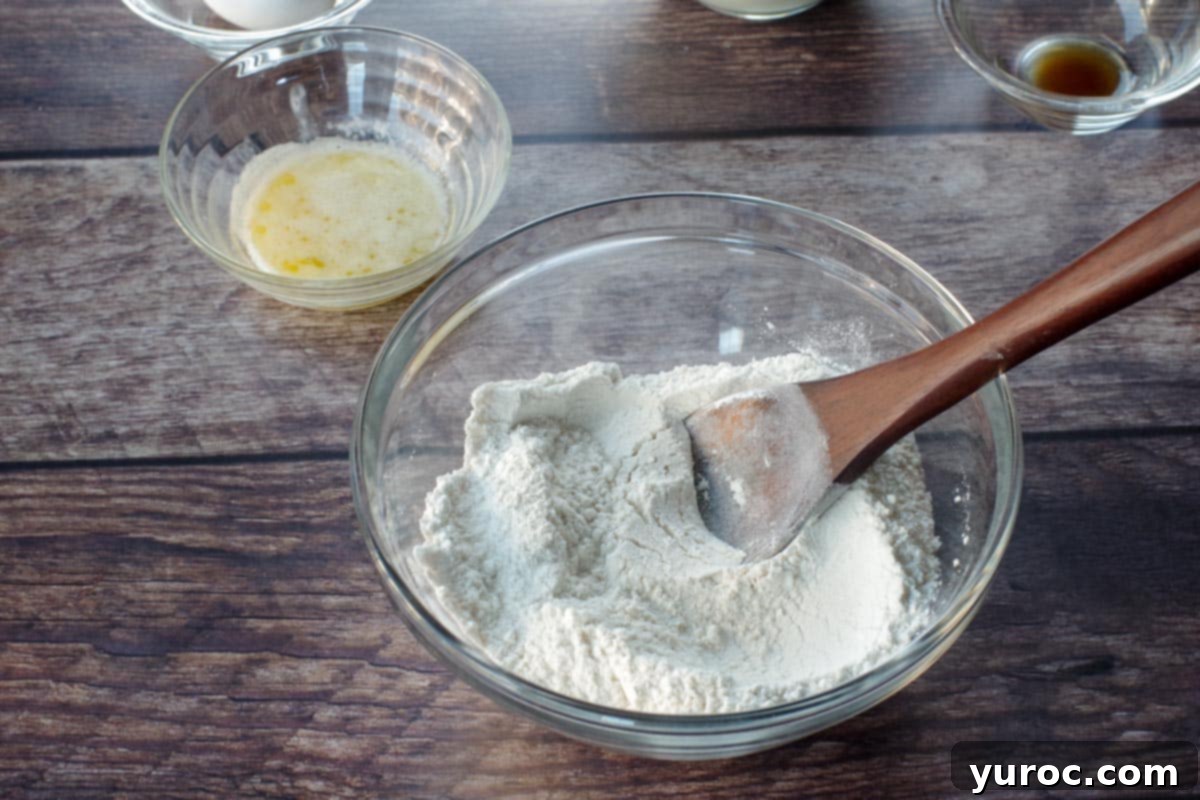Dry ingredients (flour, baking powder, salt, sugar) mixed together in a clear glass bowl with a wooden spoon.