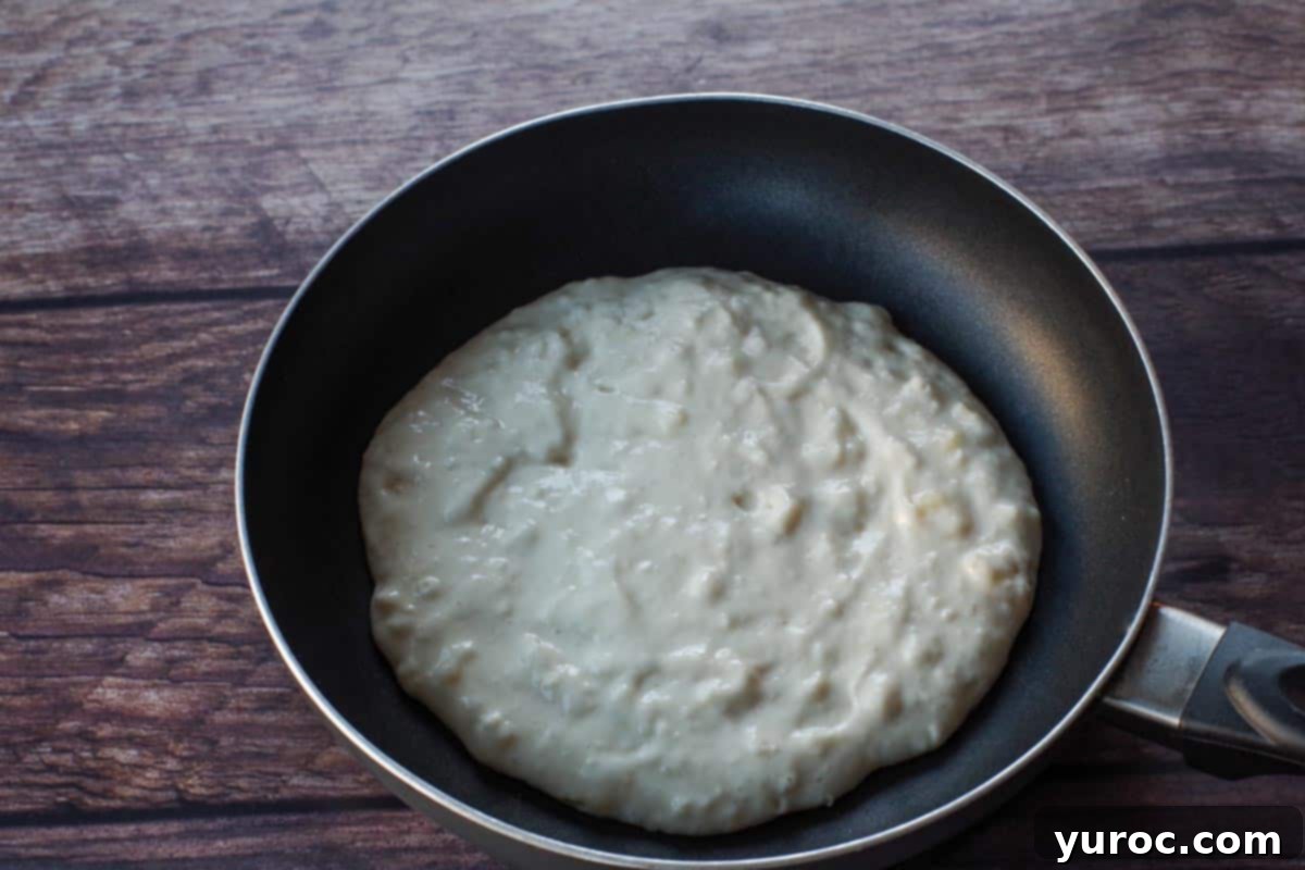 A ladle of pancake batter poured onto a hot griddle, starting to spread into a circular shape.