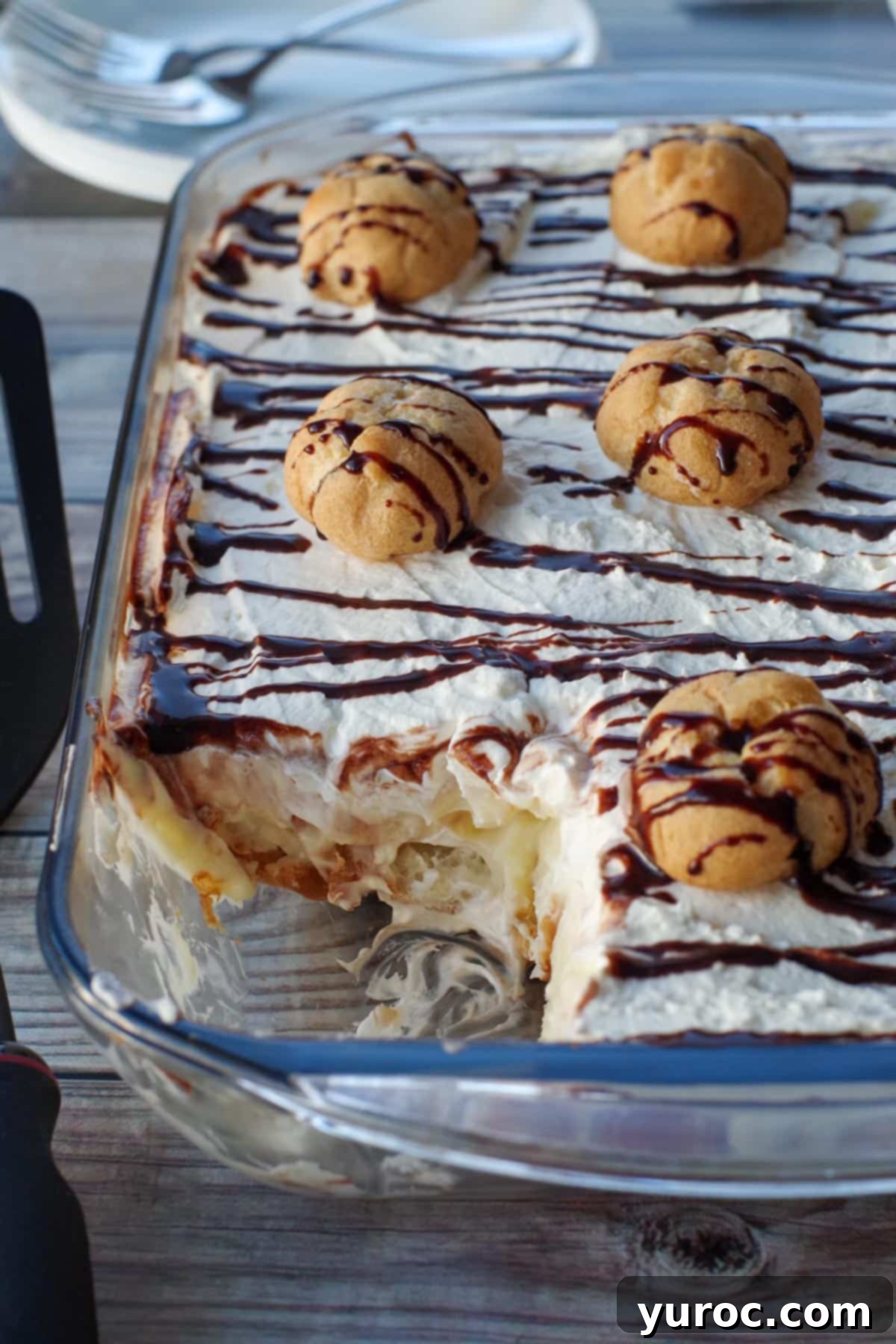 A large No-Bake Cream Puff Dessert with a slice removed, showcasing its delicious layers. Drizzled with chocolate syrup and garnished with small cream puffs.
