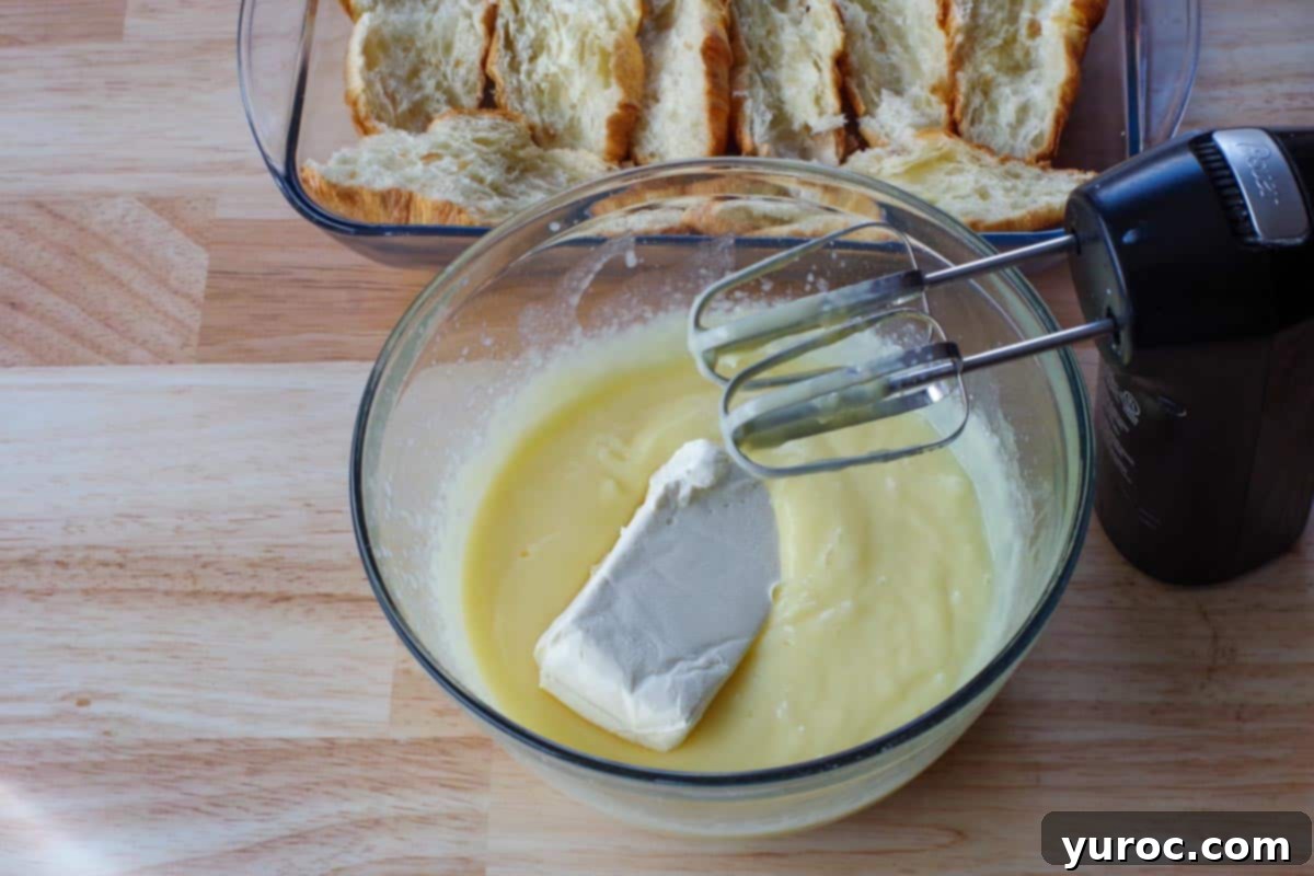 Softened cream cheese being mixed into the prepared vanilla pudding mixture in a large glass bowl using an electric hand mixer.