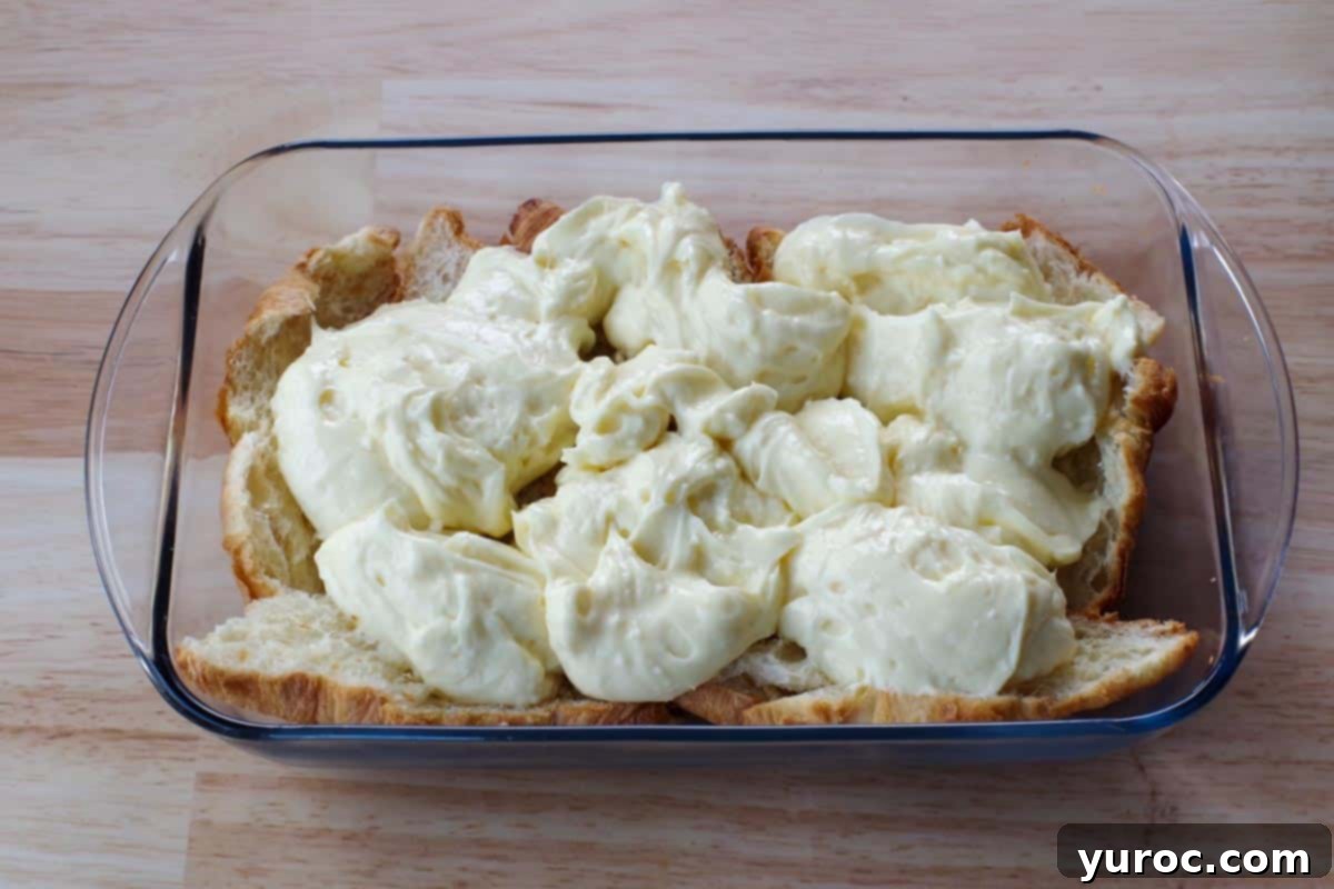 Spoonfuls of the pudding and cream cheese mixture being gently dropped onto the croissant layer in the glass dish.