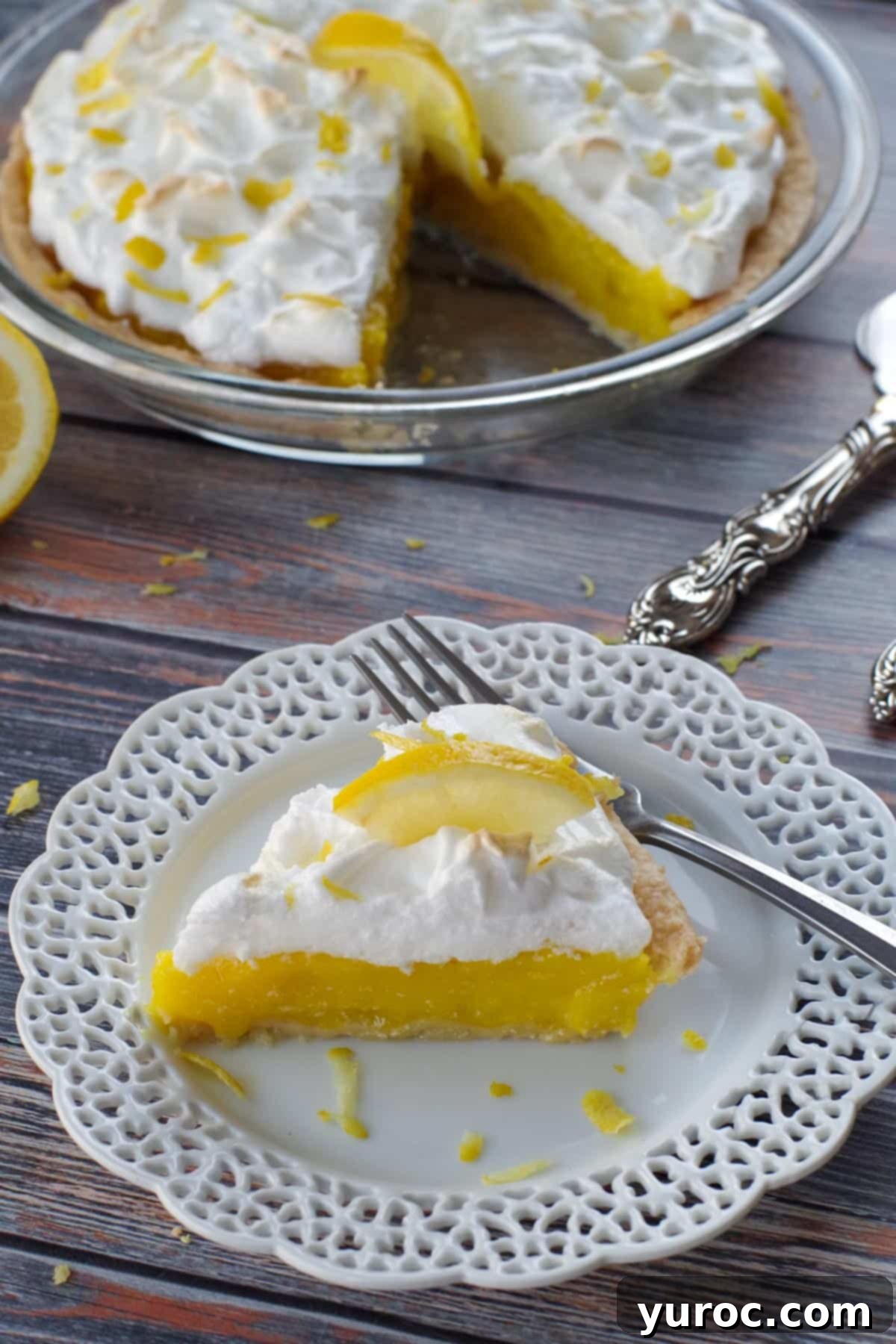 A beautifully sliced homemade lemon meringue pie on a white plate with a decorative lattice trim, with the whole pie visible in the background.