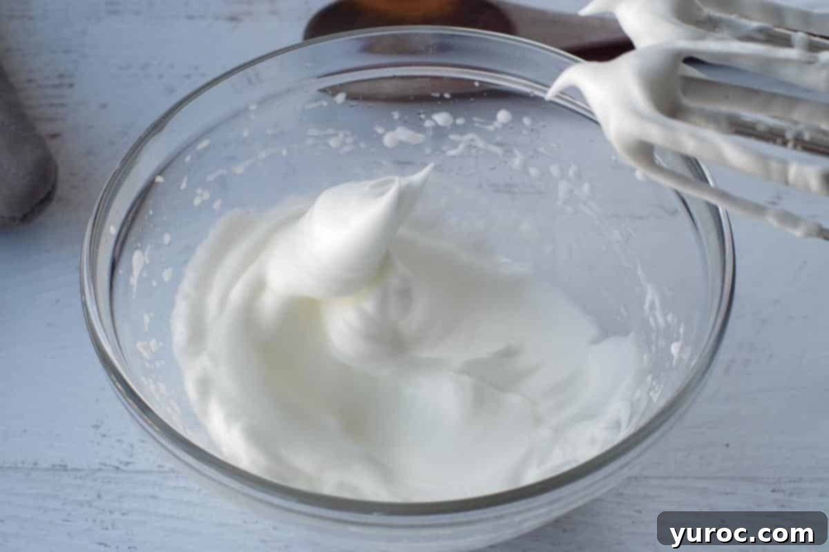 Egg whites being beaten to stiff peaks in a glass bowl with an electric mixer.