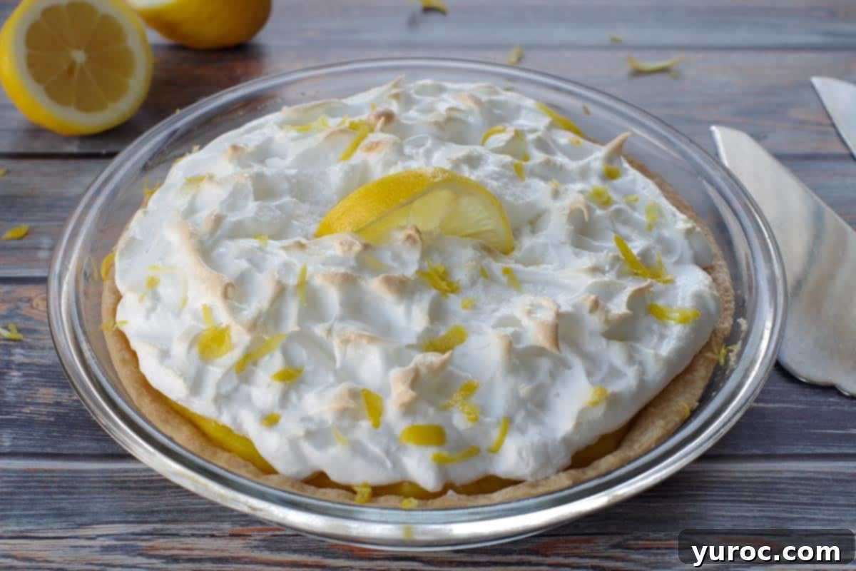 A complete old fashioned lemon meringue pie with a fresh lemon cut in half in the background, along with serving utensils.