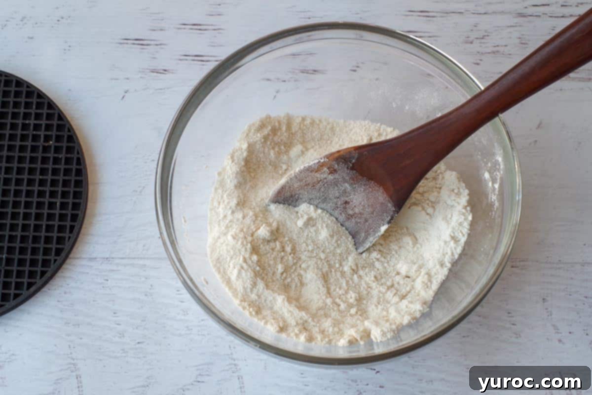Dry ingredients, flour and sugar, mixed together in a clear glass mixing bowl with a wooden spoon.