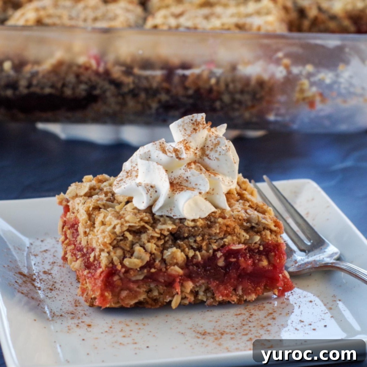 Sweet & Tart Rhubarb Oat Cookies 3 A square piece of Rhubarb Square dessert on a white plate, with a glass baking dish filled with more Rhubarb Squares blurred in the background.