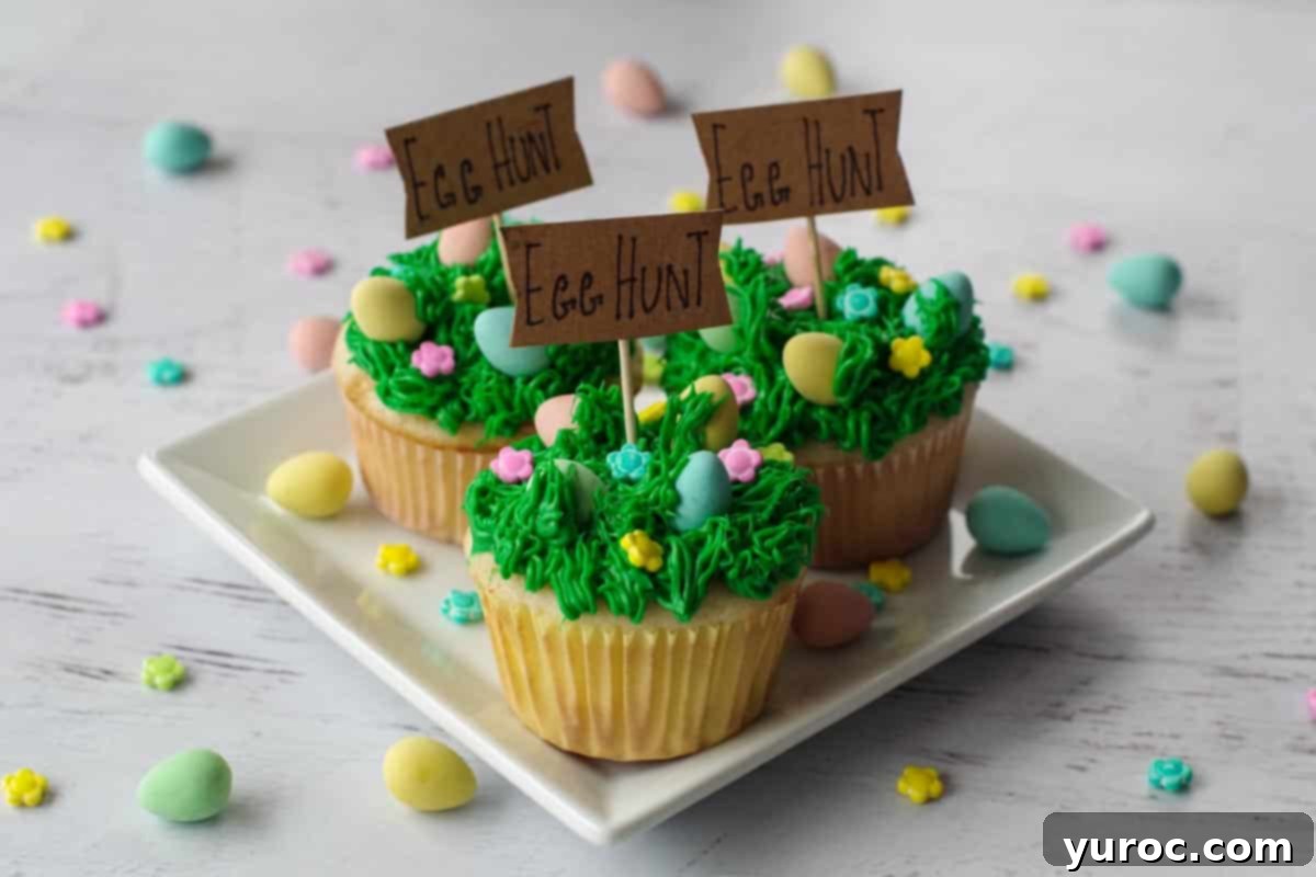 Mini Egg Hunt Cupcake Treasures 1 Three beautifully decorated Mini Egg Cupcakes arranged on a square white plate, surrounded by scattered mini eggs and delicate candy flowers, evoking an Easter theme.