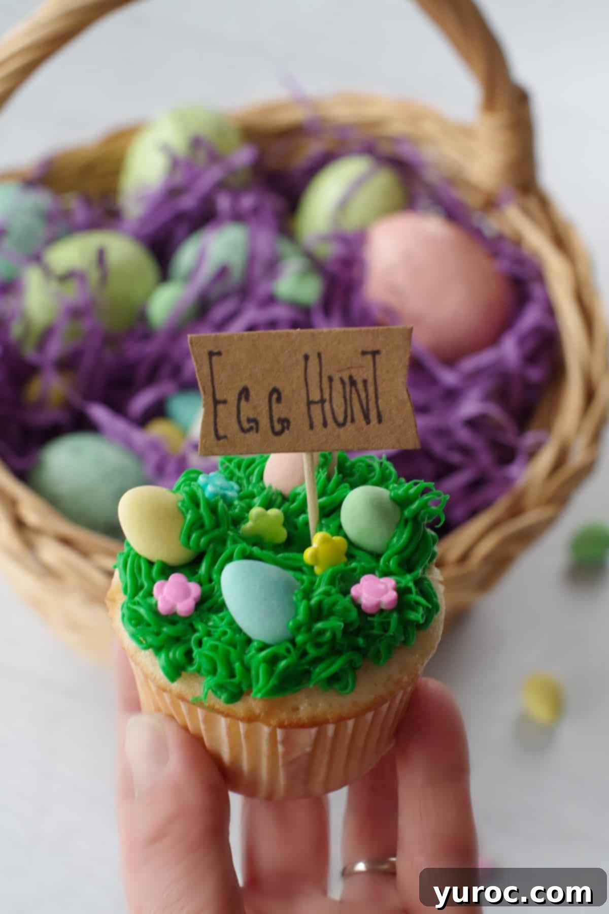 Mini Egg Hunt Cupcake Treasures 17 A vibrant Easter Mini Egg cupcake being held up against a backdrop of a colorful Easter egg basket, showcasing its festive appeal.
