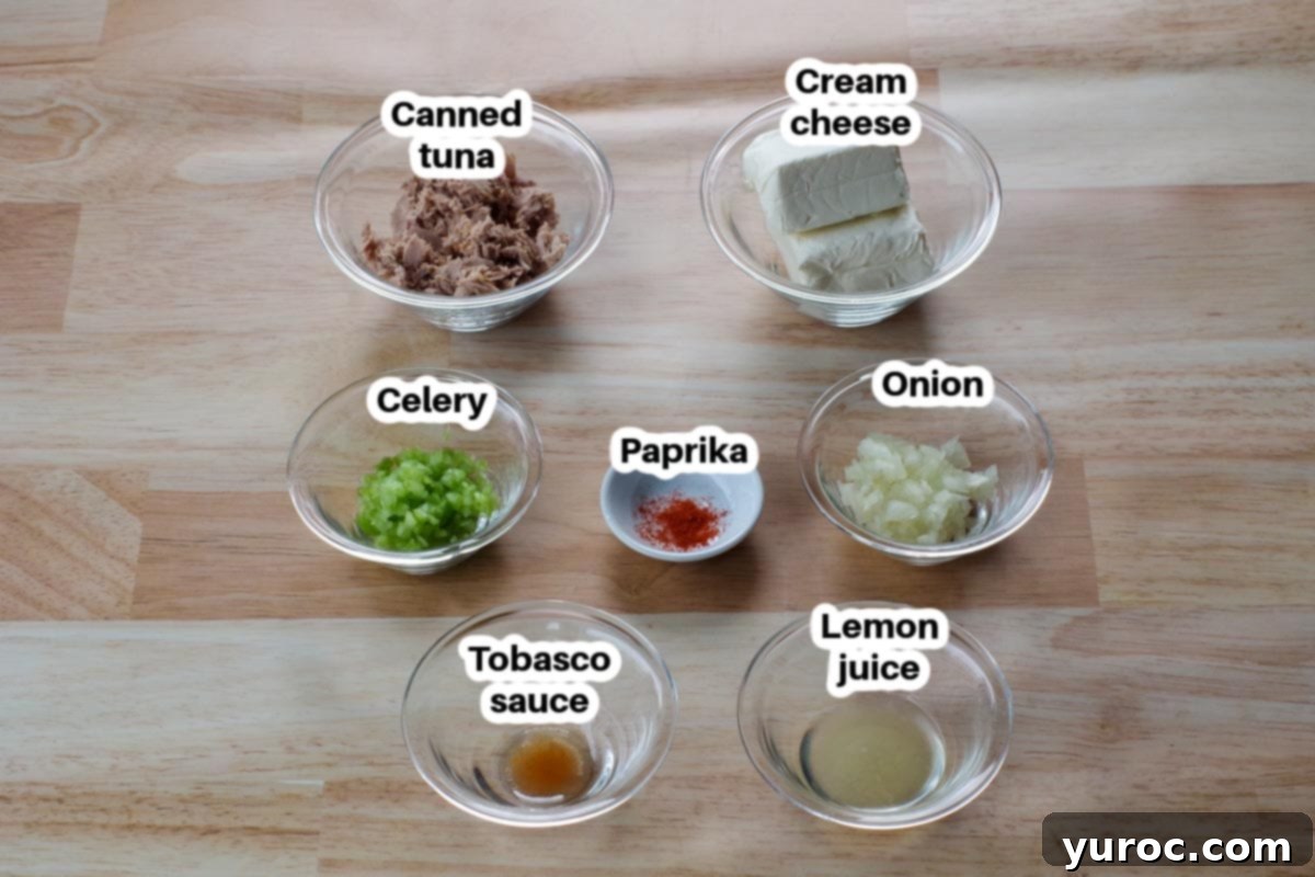 Creamy Tuna Dip 3 Individual glass bowls containing minced onion, minced celery, lemon juice, paprika, and drained canned tuna, arranged on a faux wooden surface.
