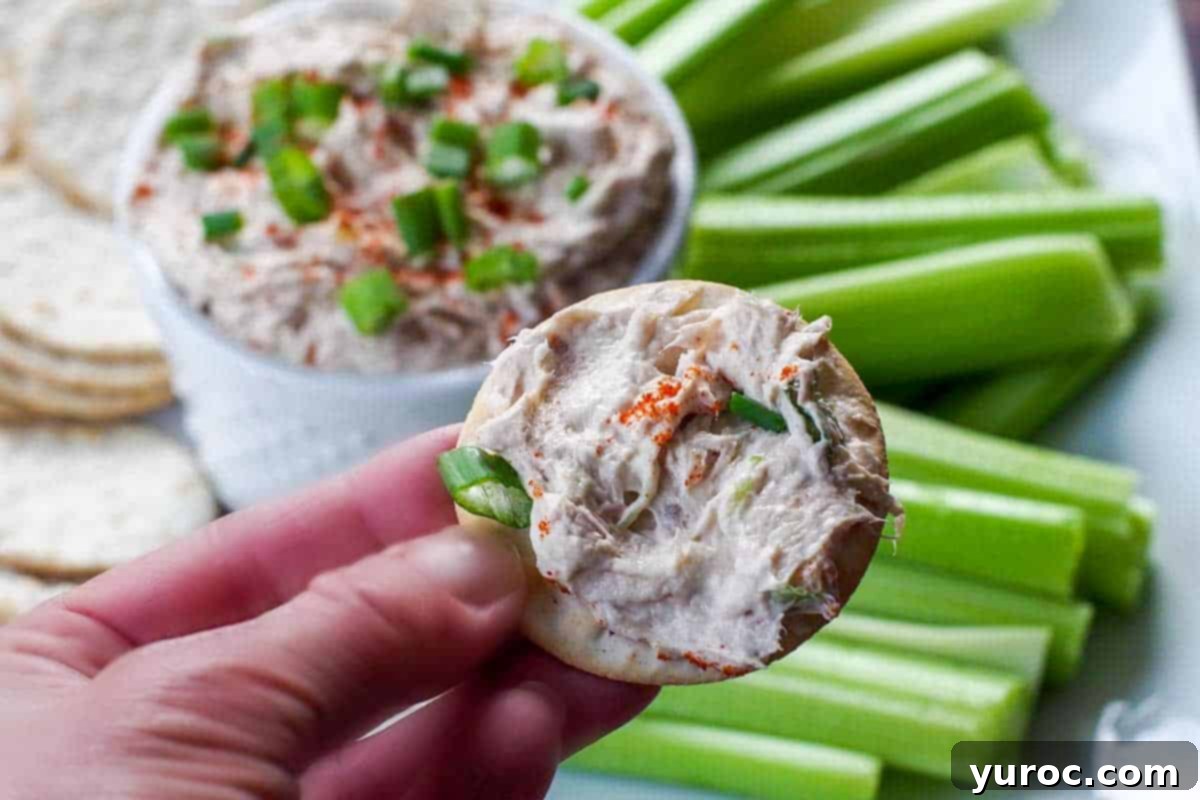Creamy Tuna Dip 7 A hand holding a cracker generously topped with tuna dip, lifted from a white serving container surrounded by more crackers and celery.