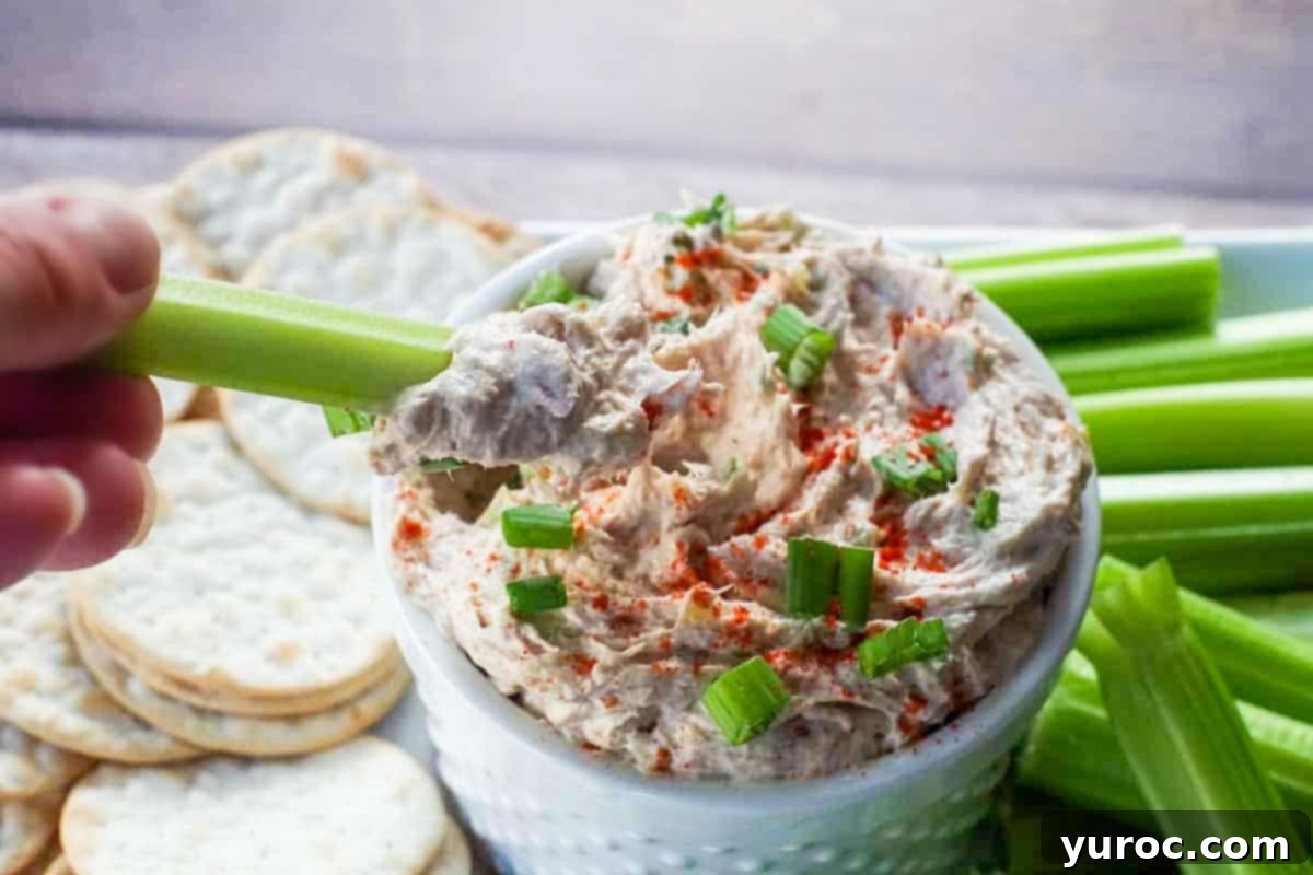Creamy Tuna Dip 8 A celery stick being dipped into the creamy tuna dip, presented in a white serving container on a tray with crackers and more celery.
