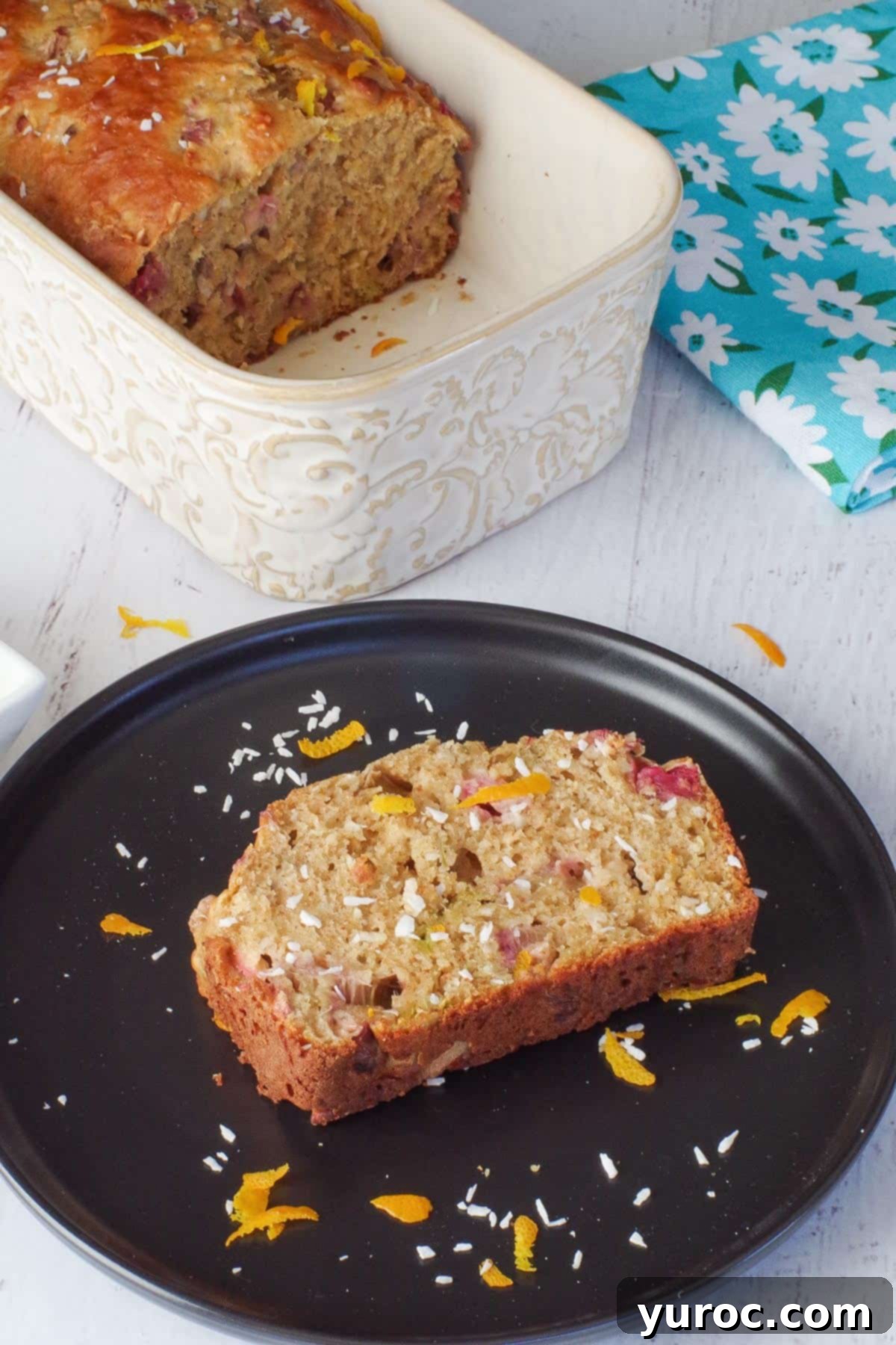 piece of healthy rhubarb bread on a black plate with a ½ a loaf of rhubarb bread in a pan in the back