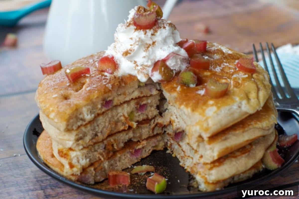 Four golden-brown rhubarb pancakes stacked high with a piece cut out, revealing the soft, fluffy interior and rhubarb pieces, topped with whipped cream and a sprinkle of cinnamon on a dark plate.