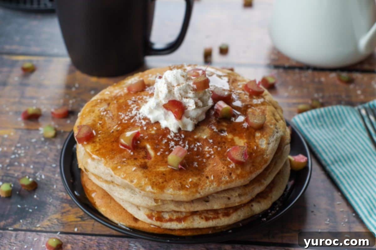 Four rhubarb pancakes stacked on a black plate, garnished with whipped cream, toasted coconut flakes, a sprinkle of cinnamon, and a drizzle of syrup. A black coffee cup and white syrup dispenser are in the background.