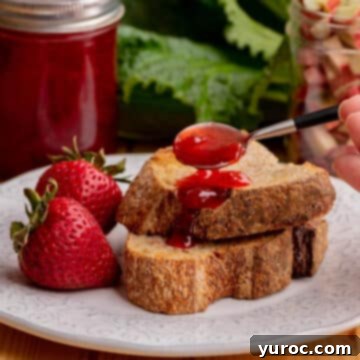 rhubarb jam on a spoon being spread over toast, on a white plate, with strawberries and container of jam in the background