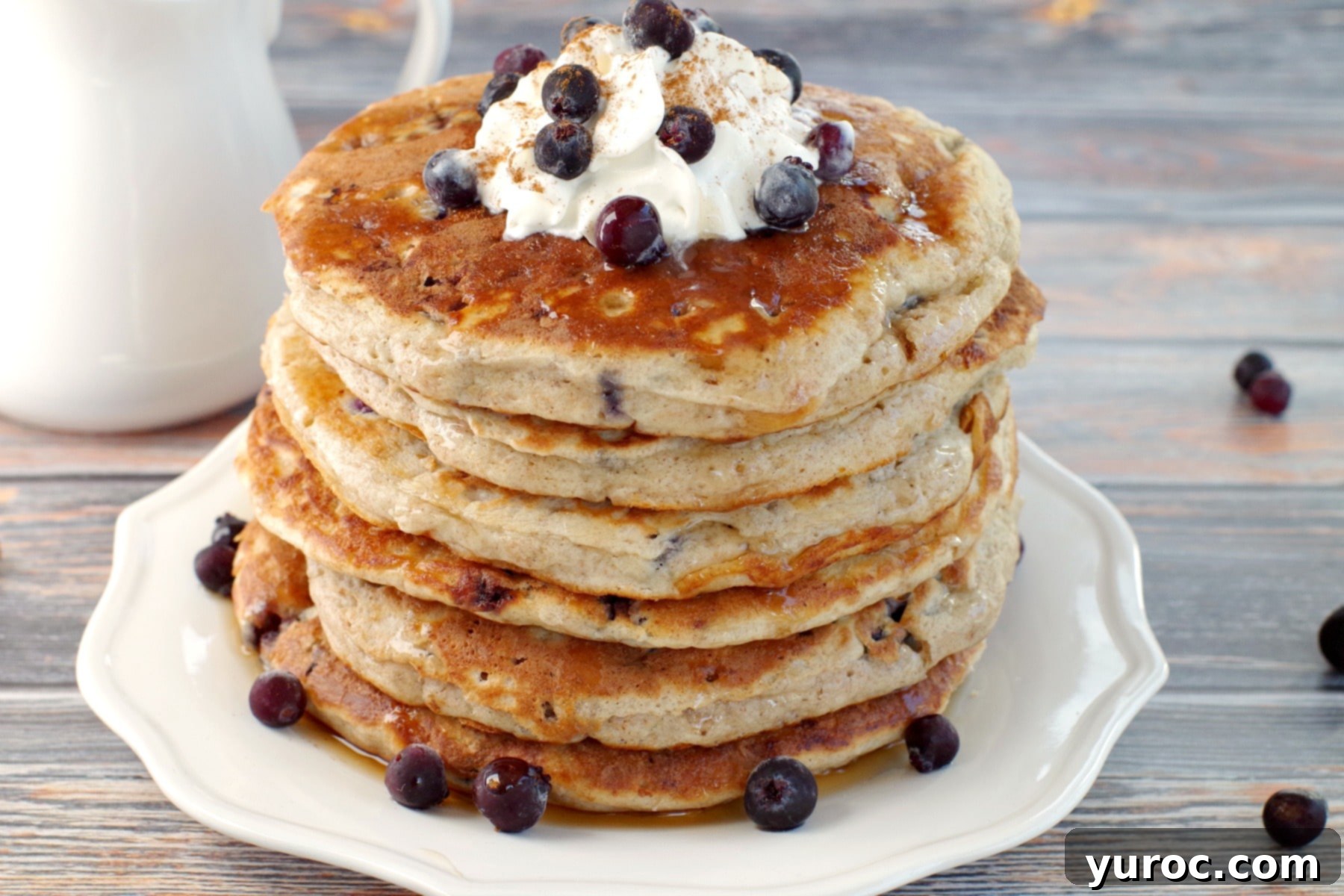 A tall stack of fluffy Saskatoon Berry Pancakes adorned with a generous dollop of whipped cream and a scattering of fresh berries on top.