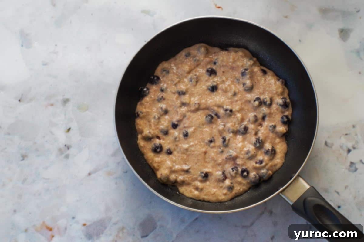 Saskatoon berry pancake batter being carefully poured into an 8-inch non-stick frying pan, preheated on a stovetop.