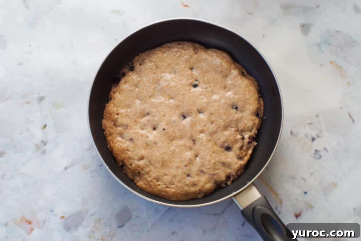 A Saskatoon berry pancake sizzling in a pan, with visible bubbles forming on its surface, indicating it's ready to be flipped.