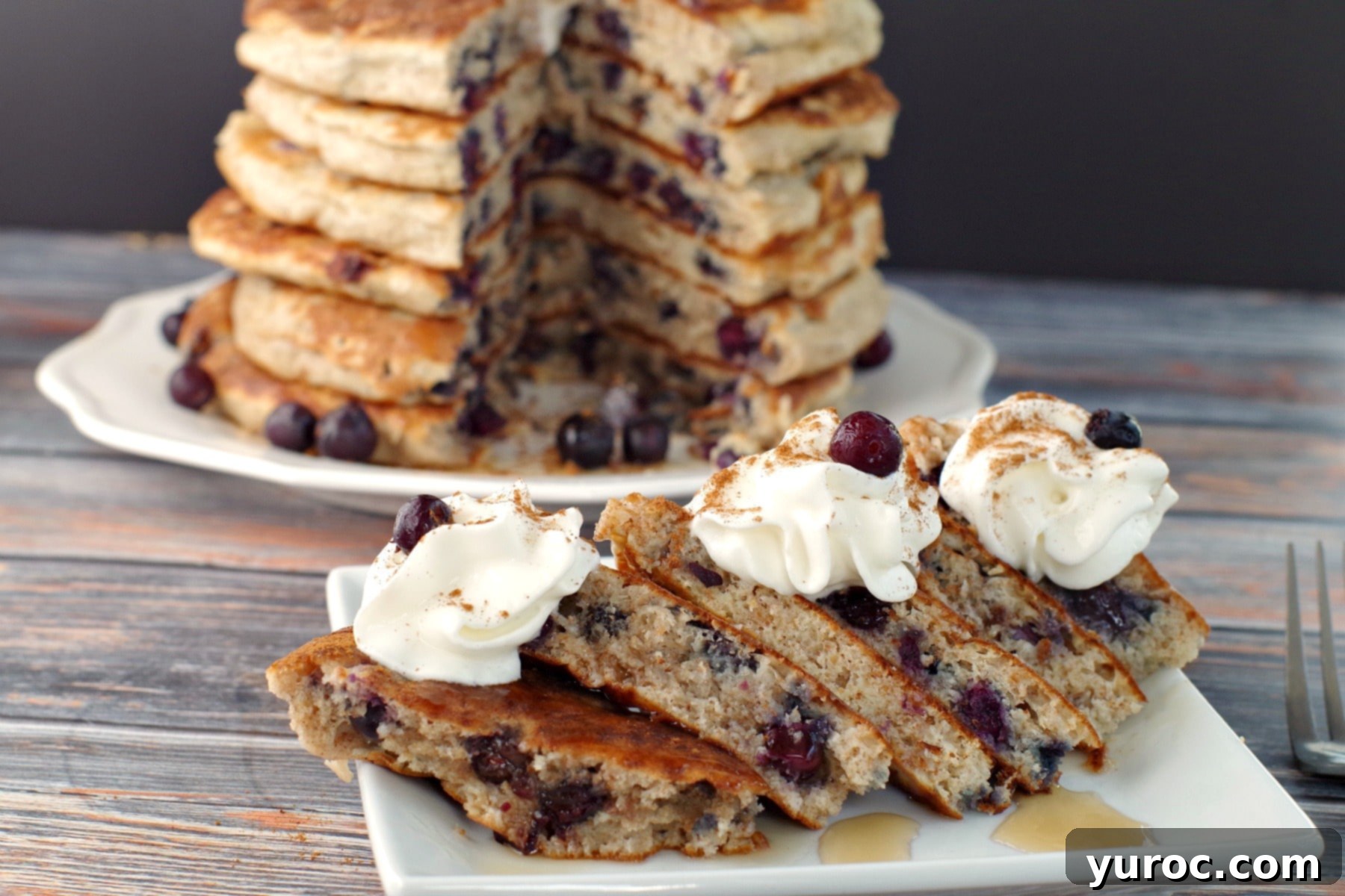 A single, perfectly cut out Saskatoon berry pancake, showcasing its fluffy interior and visible berries, sits on a white plate with a full stack of pancakes blurred in the background.