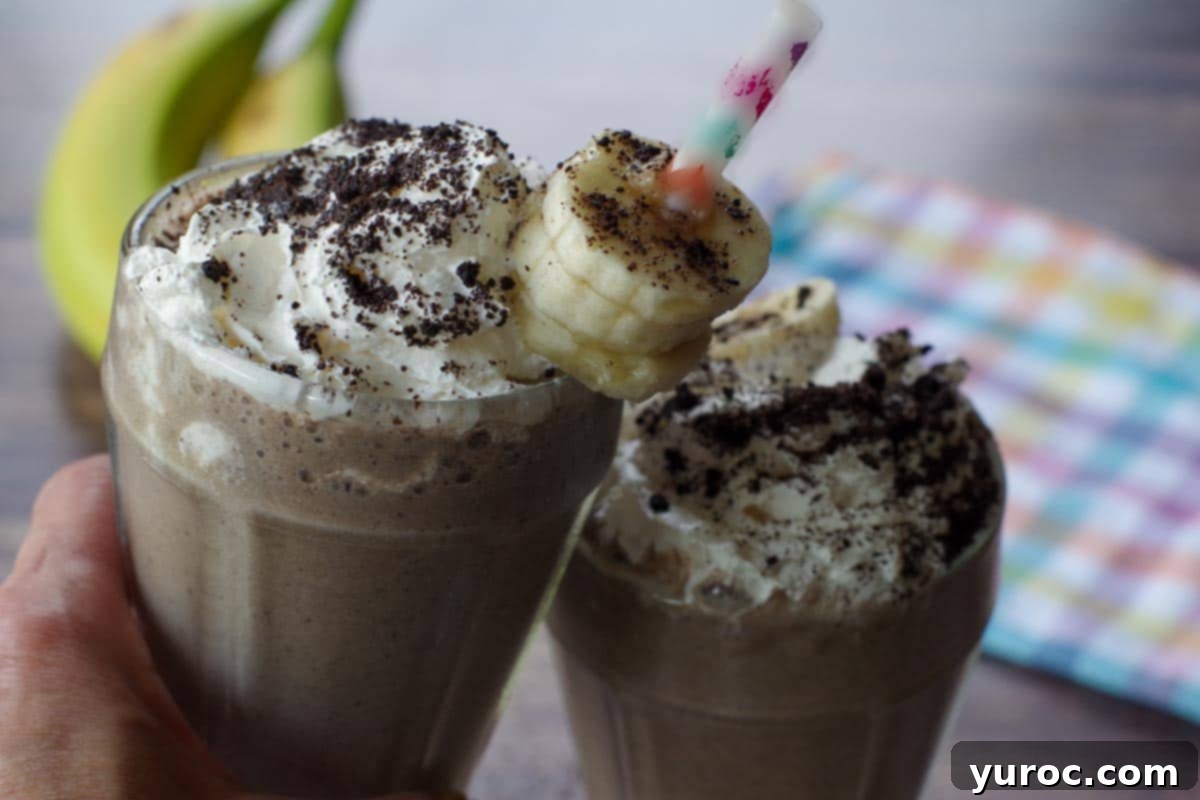 Guilt-Free Oreo Banana Milkshake 2 Two glasses of Healthy Oreo Banana Milkshake clinking together, with fresh bananas in the soft-focus background.