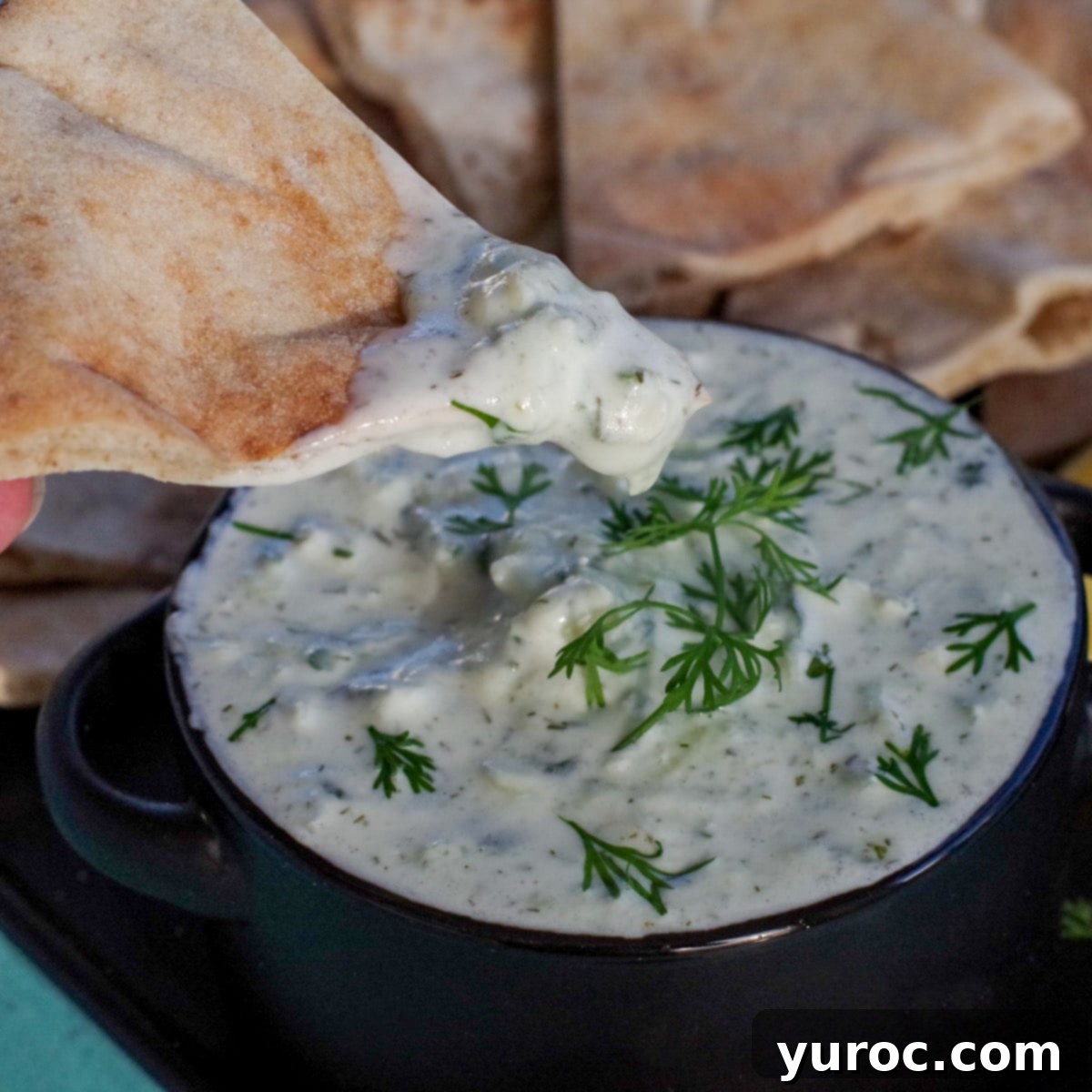 triangle of pita bread being dipped into tzatziki sauce in black bowl on black tray with pita bread in background and lemon wedges