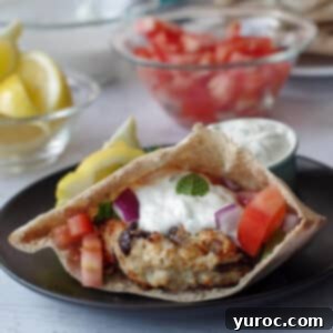 Greek chicken burger in a pita on a black plate with lemon wedge and small dish of tzatziki
