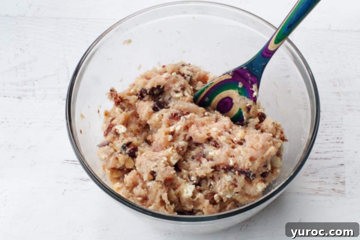 Greek chicken burger ingredients mixed together in large glass bowl with colorful wooden sppon
